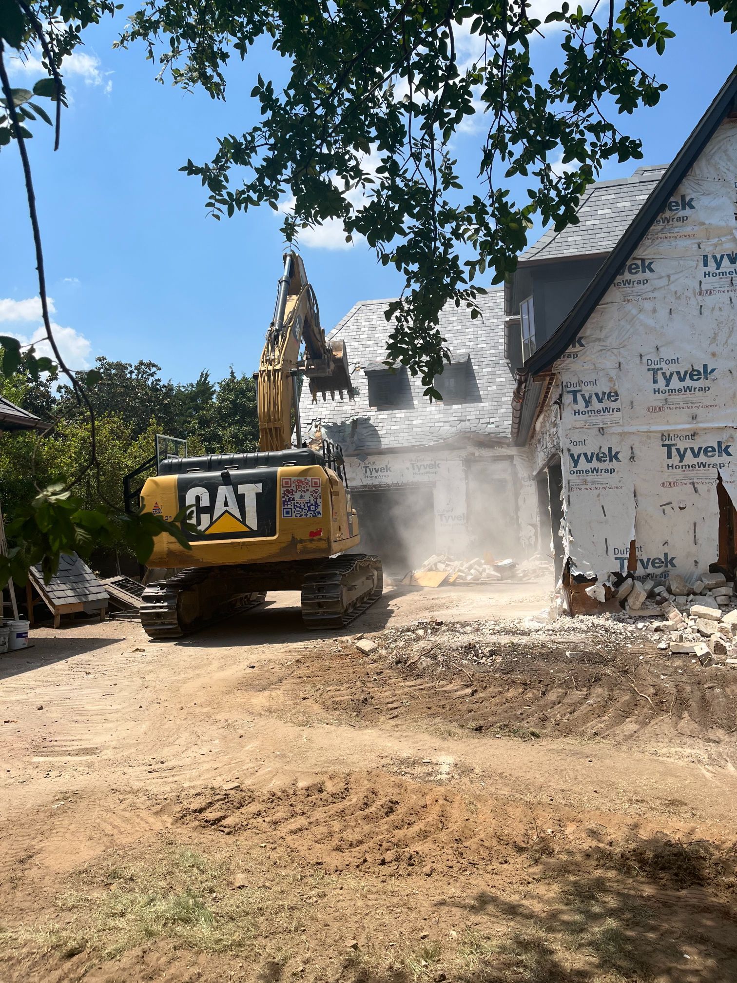 Yellow CAT excavator demolishing a house wrapped in Tyvek on a sunny construction site