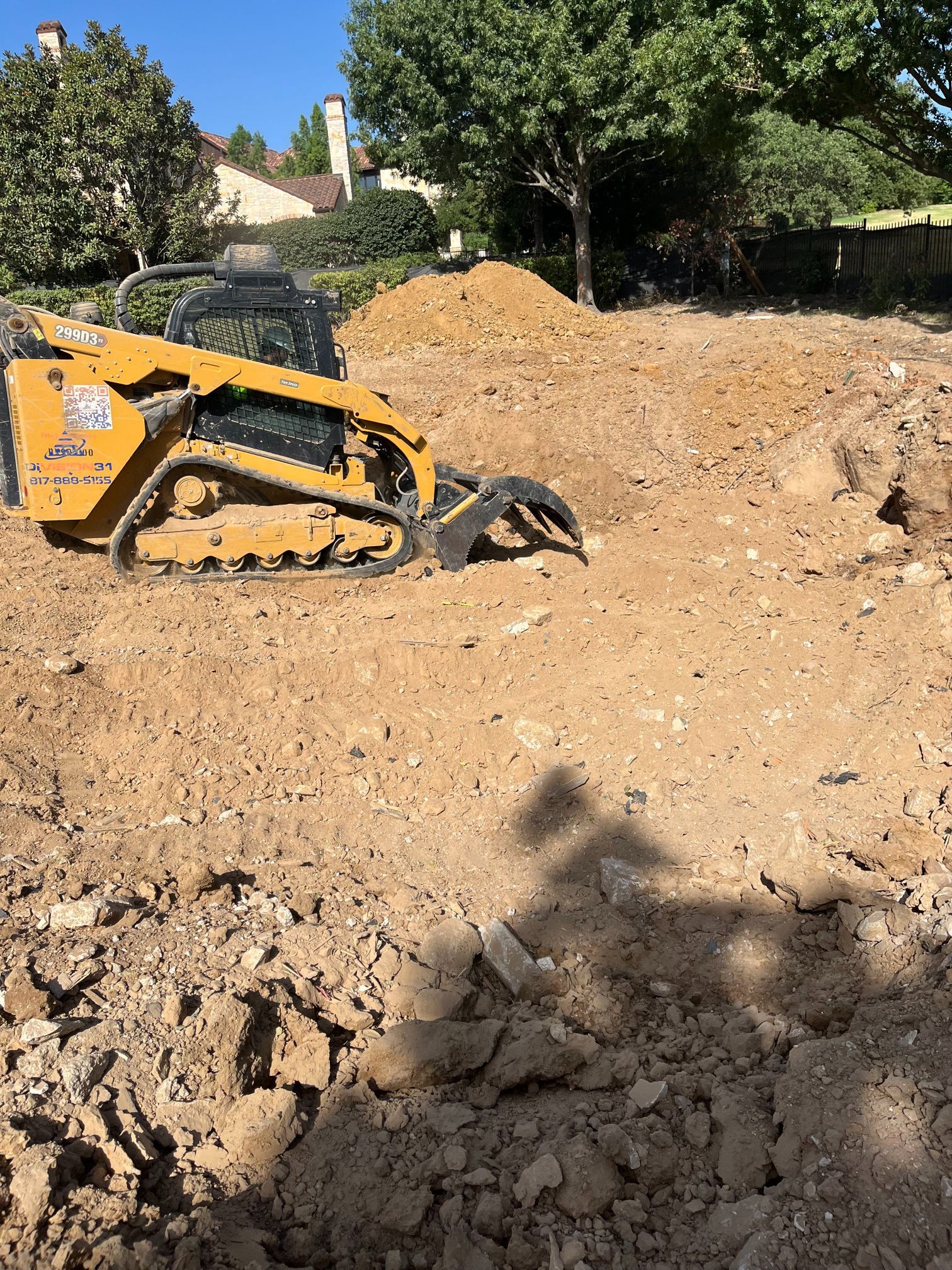 Yellow skid-steer loader on a dirt construction site, moving soil and rocks near trees and houses.