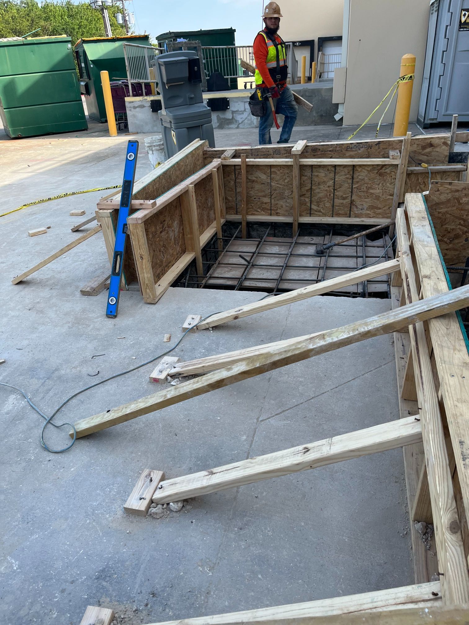 Construction site with open trench, exposed rebar, wooden formwork, and a worker in a safety vest nearby