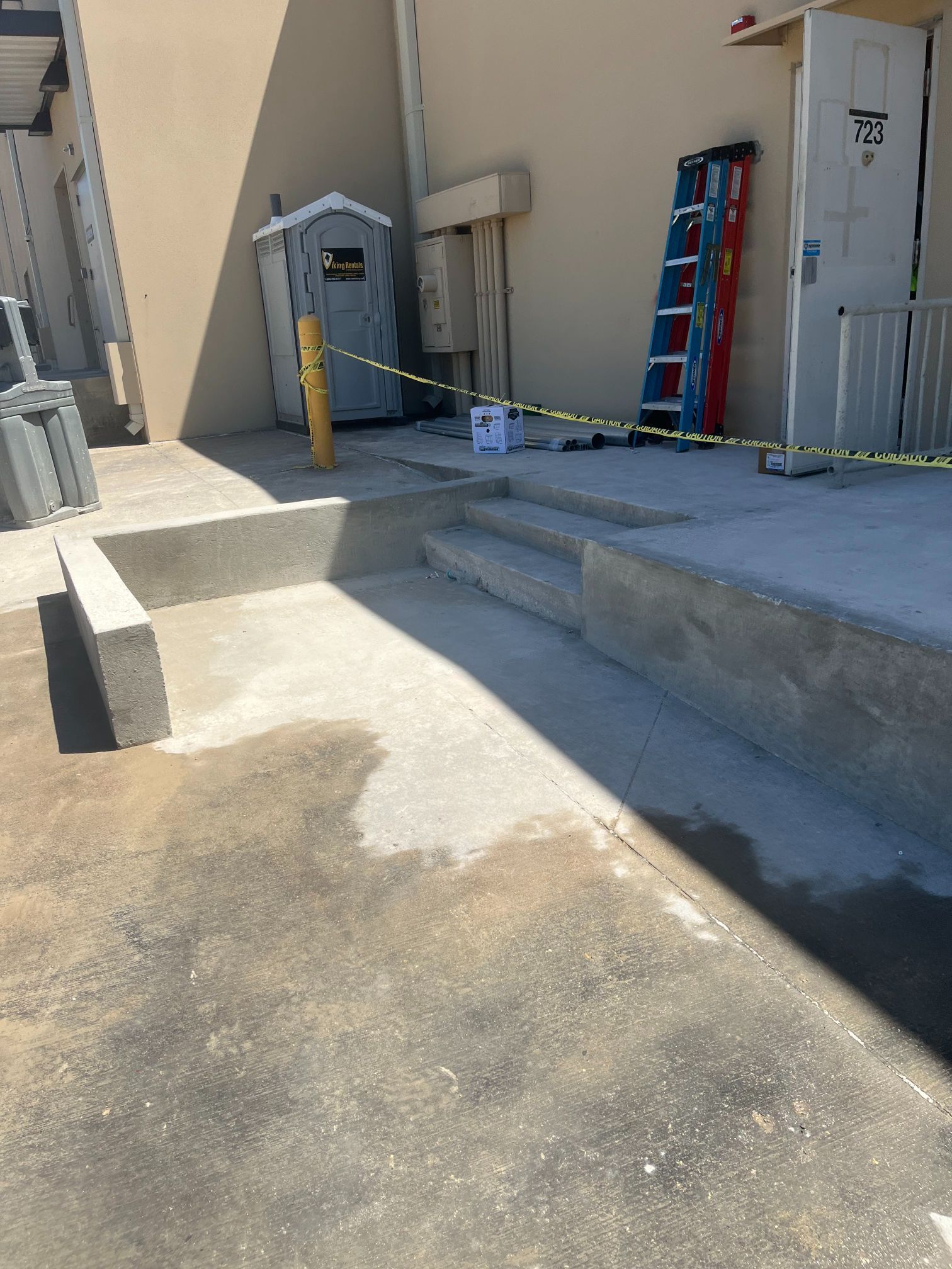 Concrete ramp area with yellow caution tape, a ladder, and utility doors beside a building