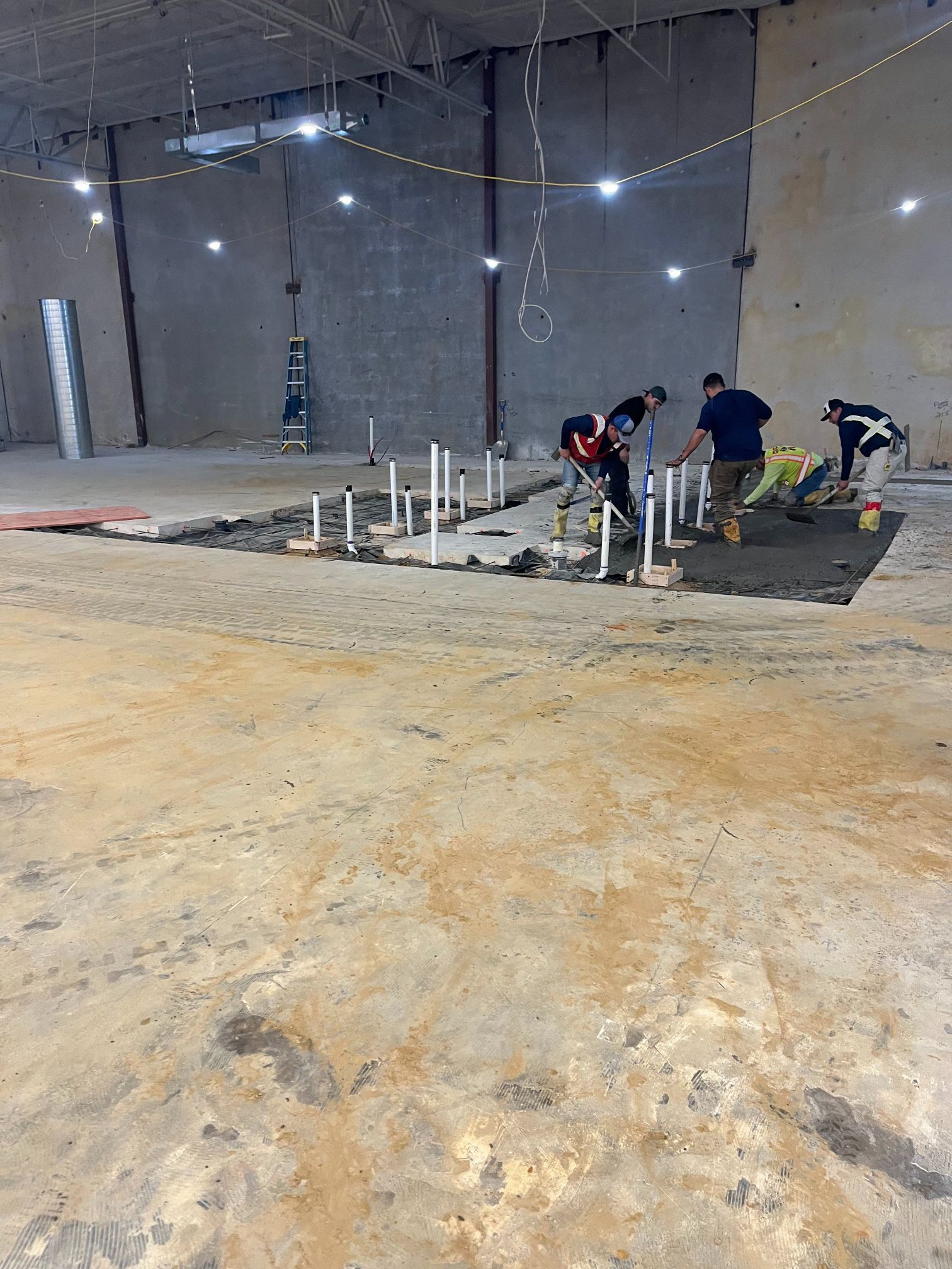 Workers pouring concrete around pipe stubs in an unfinished indoor construction site