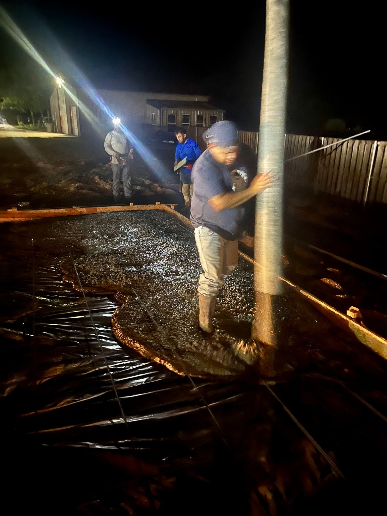 Workers pouring concrete on a dark construction site at night, with one smoothing around a vertical pole.