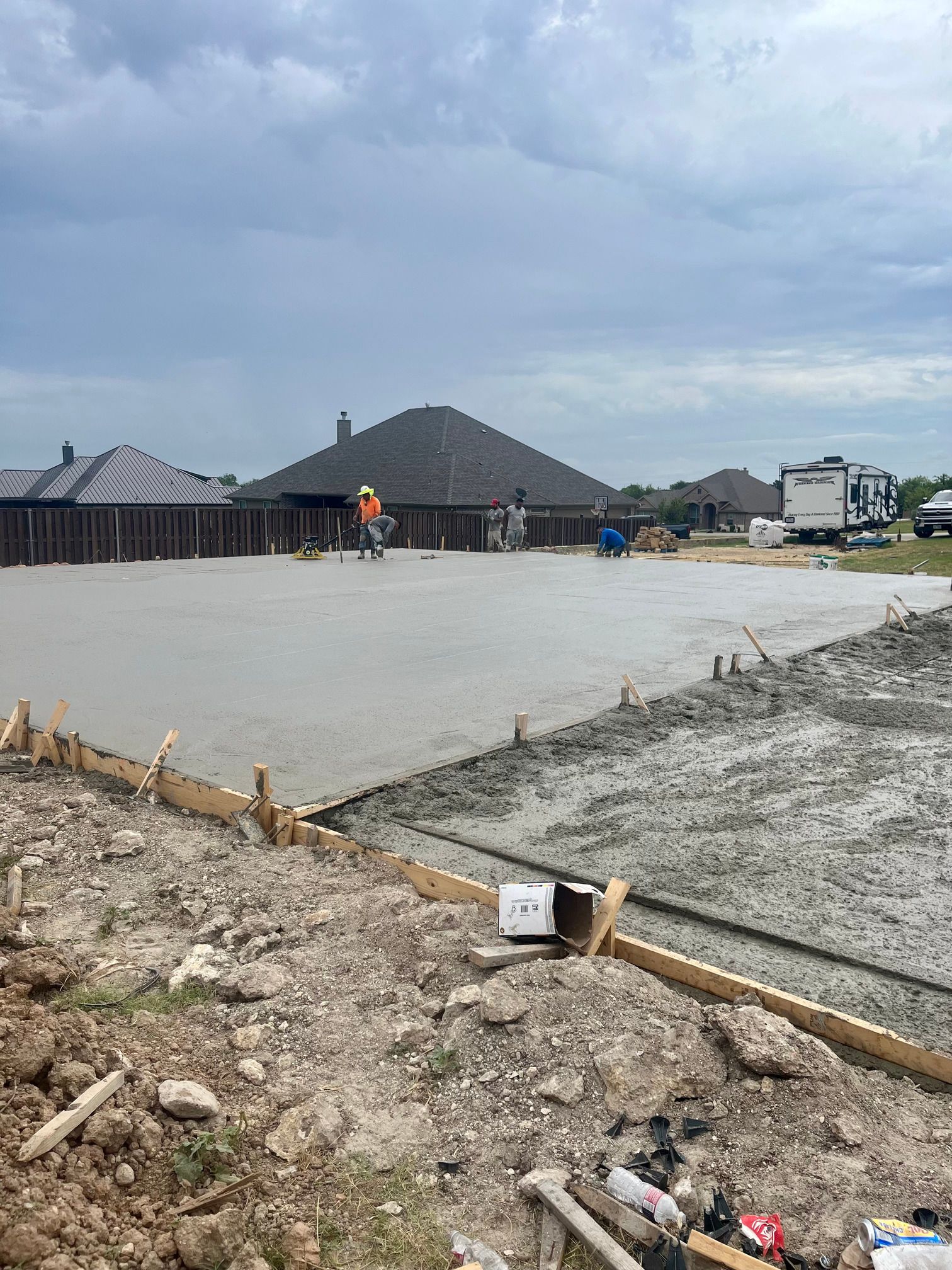 Concrete slab construction site with workers, wooden forms, and houses under a cloudy sky