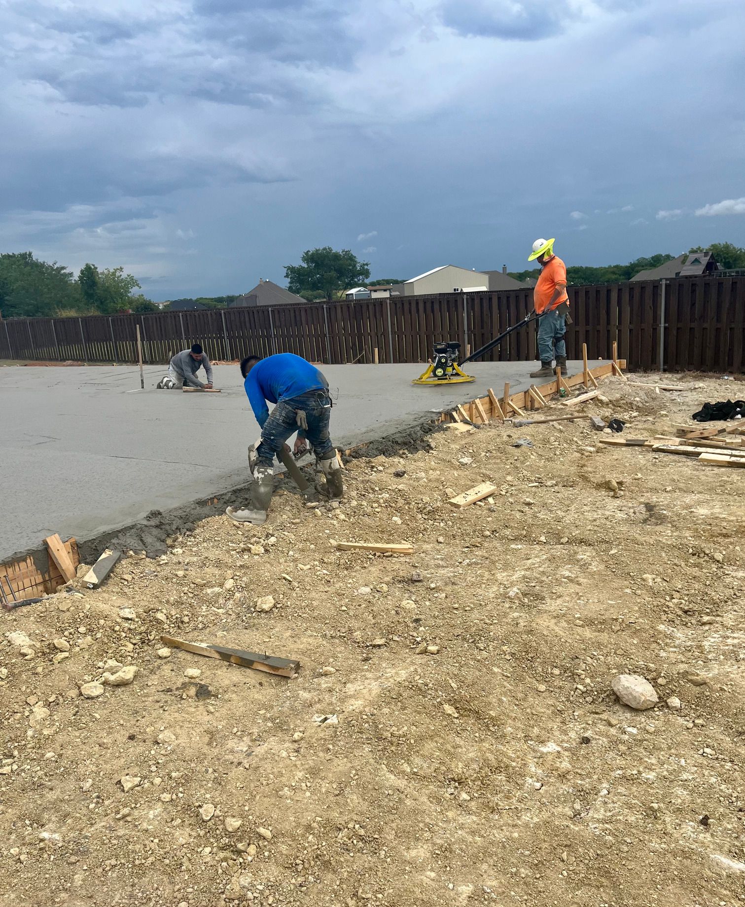 Workers pouring and smoothing concrete at a construction site under a cloudy sky.