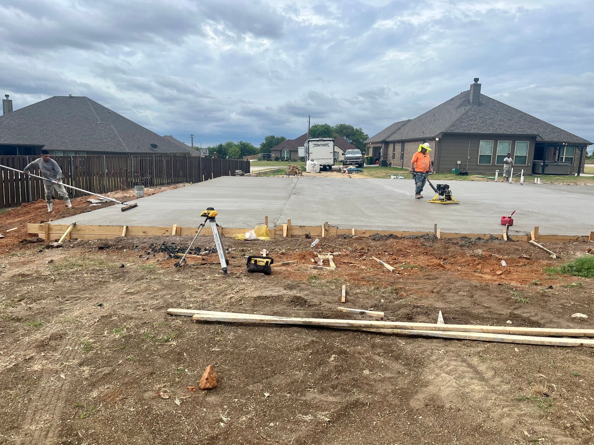 Construction site with concrete slab, workers, and unfinished suburban houses under cloudy sky