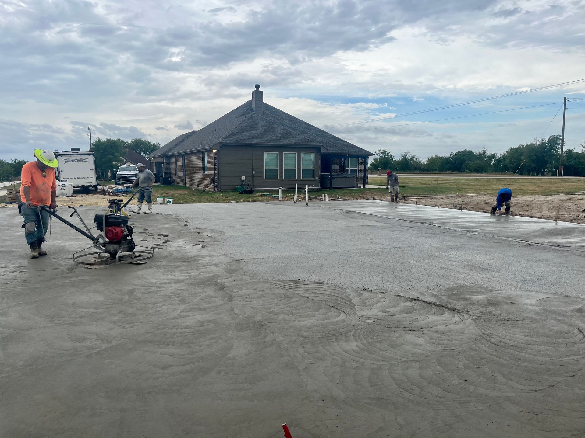 Workers pouring and smoothing concrete in front of a small building under a cloudy sky