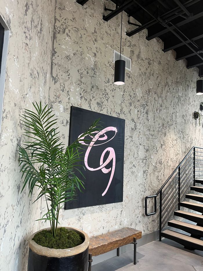 Interior staircase with textured stone wall, black sign with pink logo, potted palm, and bench