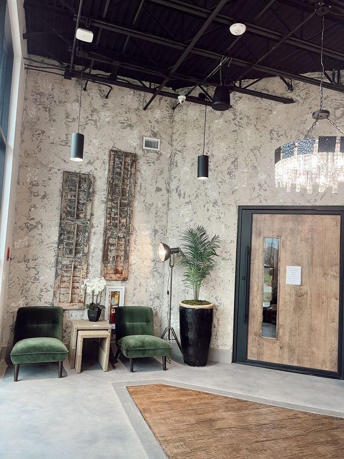 Modern lounge with black chairs, green stools, wood table, tall plants, and rustic textured walls under high ceilings