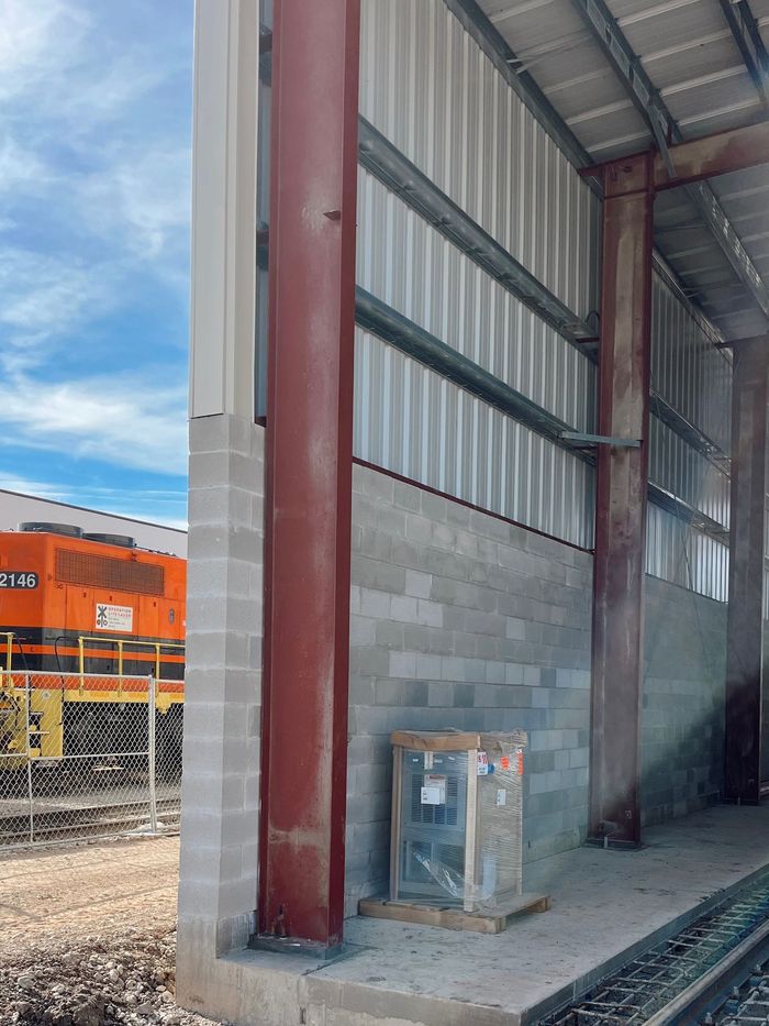 Industrial warehouse interior with red steel column, stacked cinder blocks, and an orange train outside.