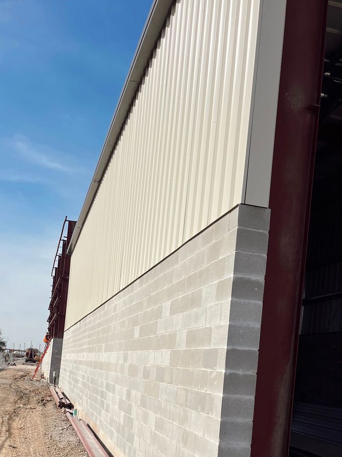 Industrial warehouse wall with beige siding, cinder blocks, and maroon trim along a dirt lot under blue sky