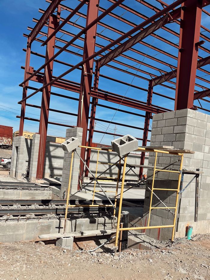 Steel building frame under construction with concrete block wall and scaffolding in front.
