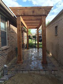 Covered brick walkway between two buildings with wooden posts and a pergola roof leading to a gate.