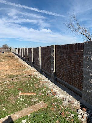 Long brick wall with concrete posts beside a grassy dirt path under a blue sky.