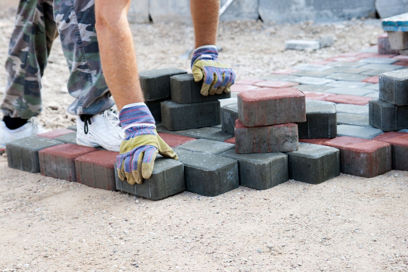 Hands in gloves laying gray paving stones beside red bricks on a sandy construction site