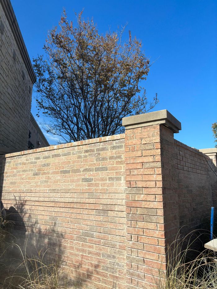 Brick wall with a square pillar beside a building, with a tree and clear blue sky in the background