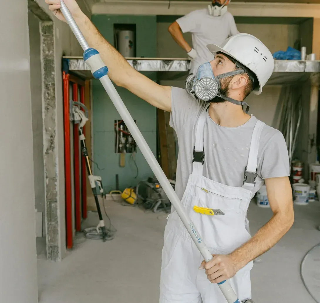 Two construction workers in hard hats and masks sand a ceiling and wall in an interior space under renovation.
