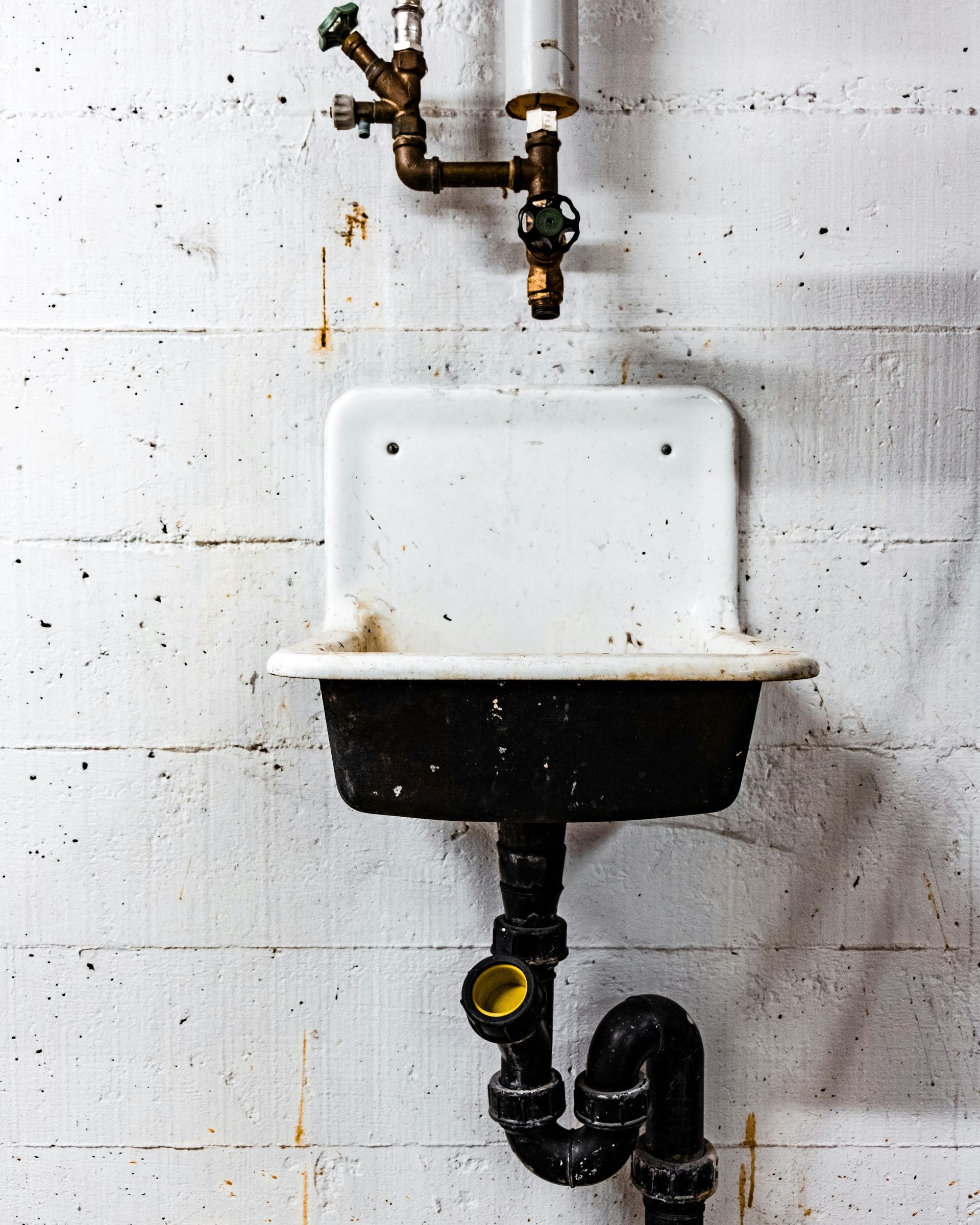 A white basin sink mounted to a white cinder block wall, with exposed brass plumbing above and a black drain pipe below.