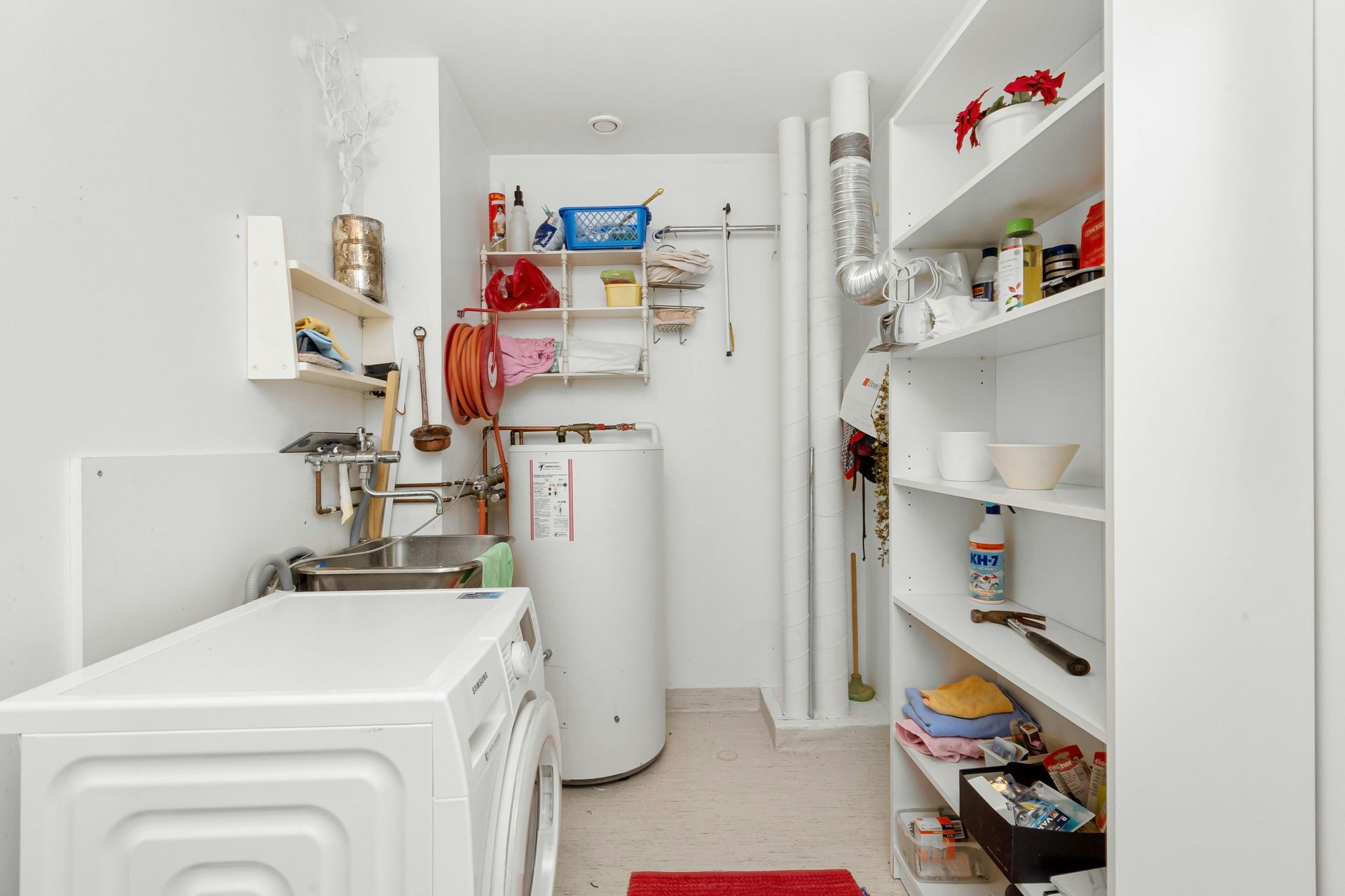 A bright utility room with a white washing machine, water heater, wall shelving, and a tall storage unit.
