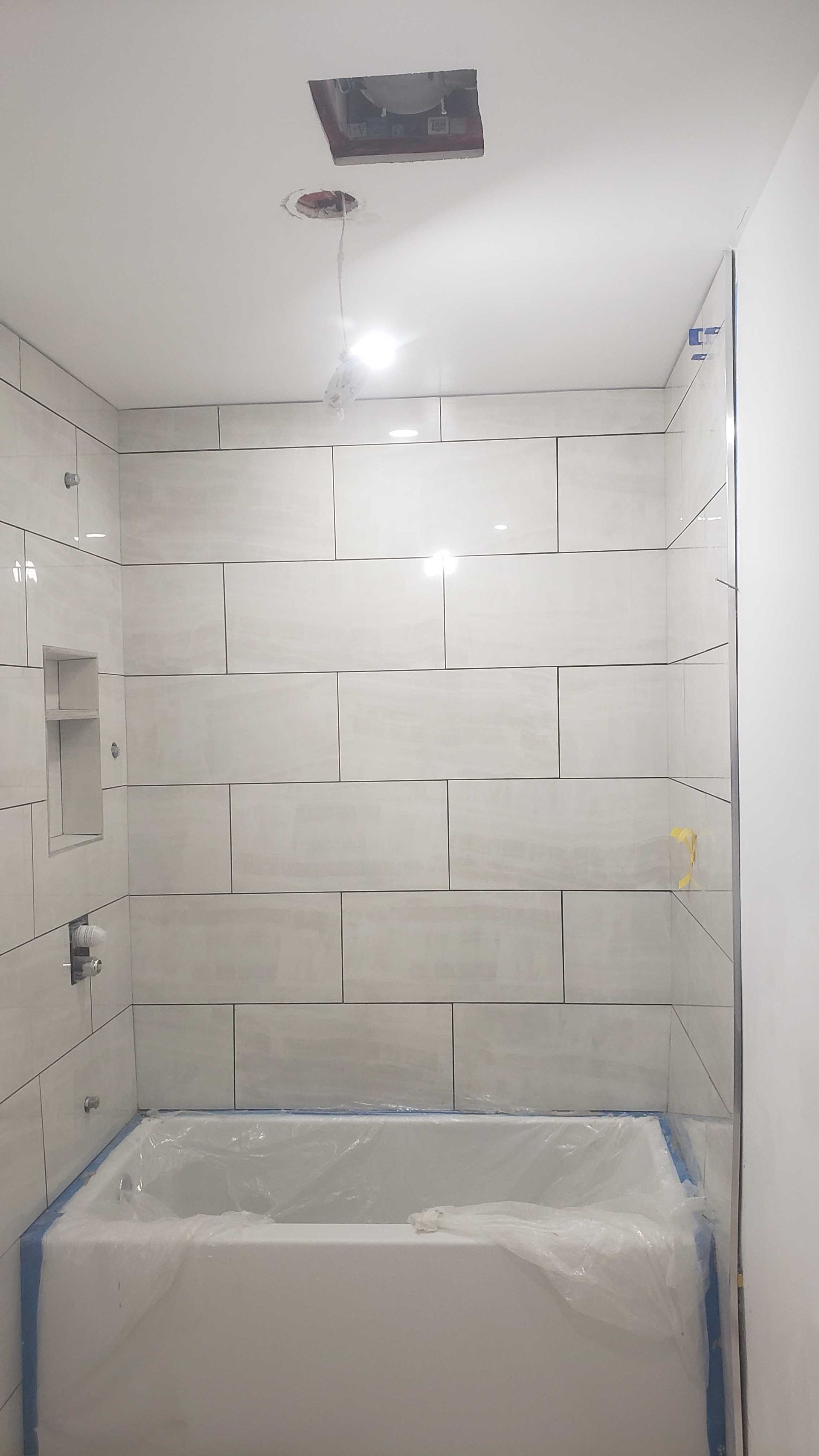 Douche inachevée avec murs carrelés blancs, éclairage au plafond et baignoire recouverte de plastique.