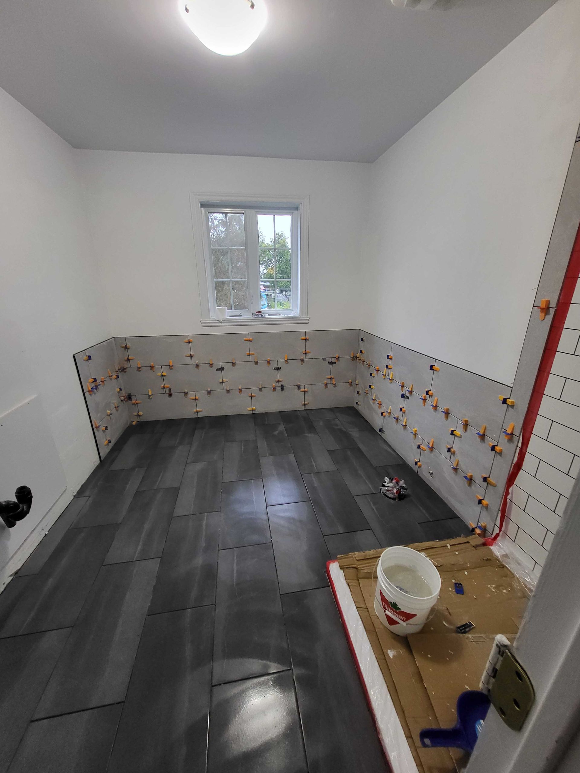 Petite salle de bains en rénovation avec sol carrelé foncé, murs à moitié carrelés, fenêtre et matériel de peinture sur un tableau.