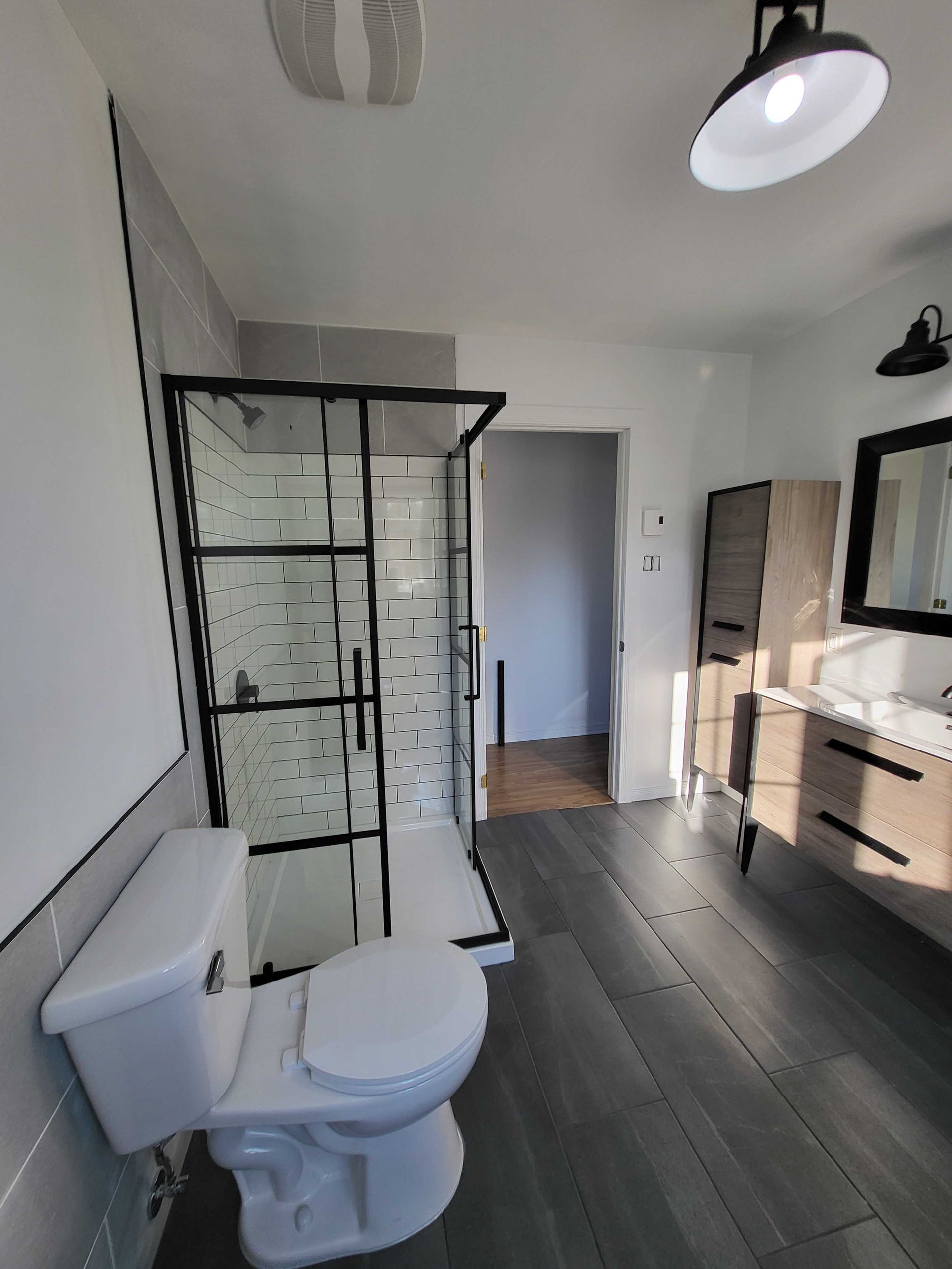 Salle de bains moderne avec toilettes, douche en verre, sol carrelé gris et meuble-lavabo sous une lumière vive.