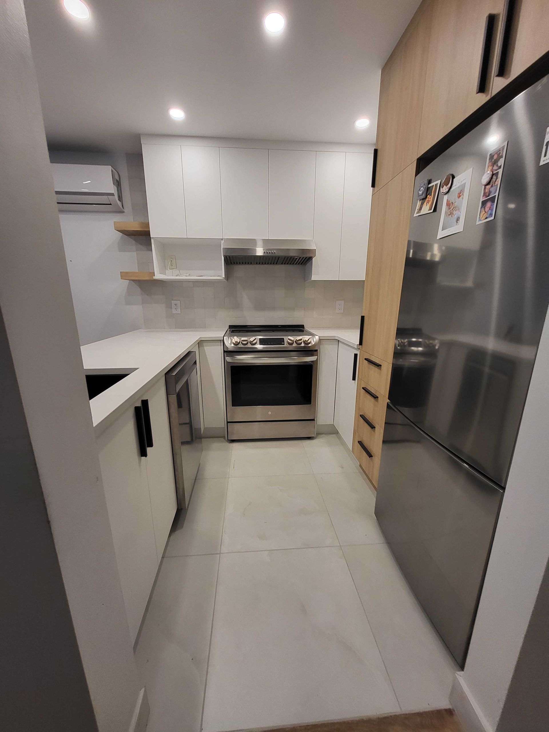 Cuisine moderne étroite avec placards blancs, appareils électroménagers en acier inoxydable et carrelage clair au sol.