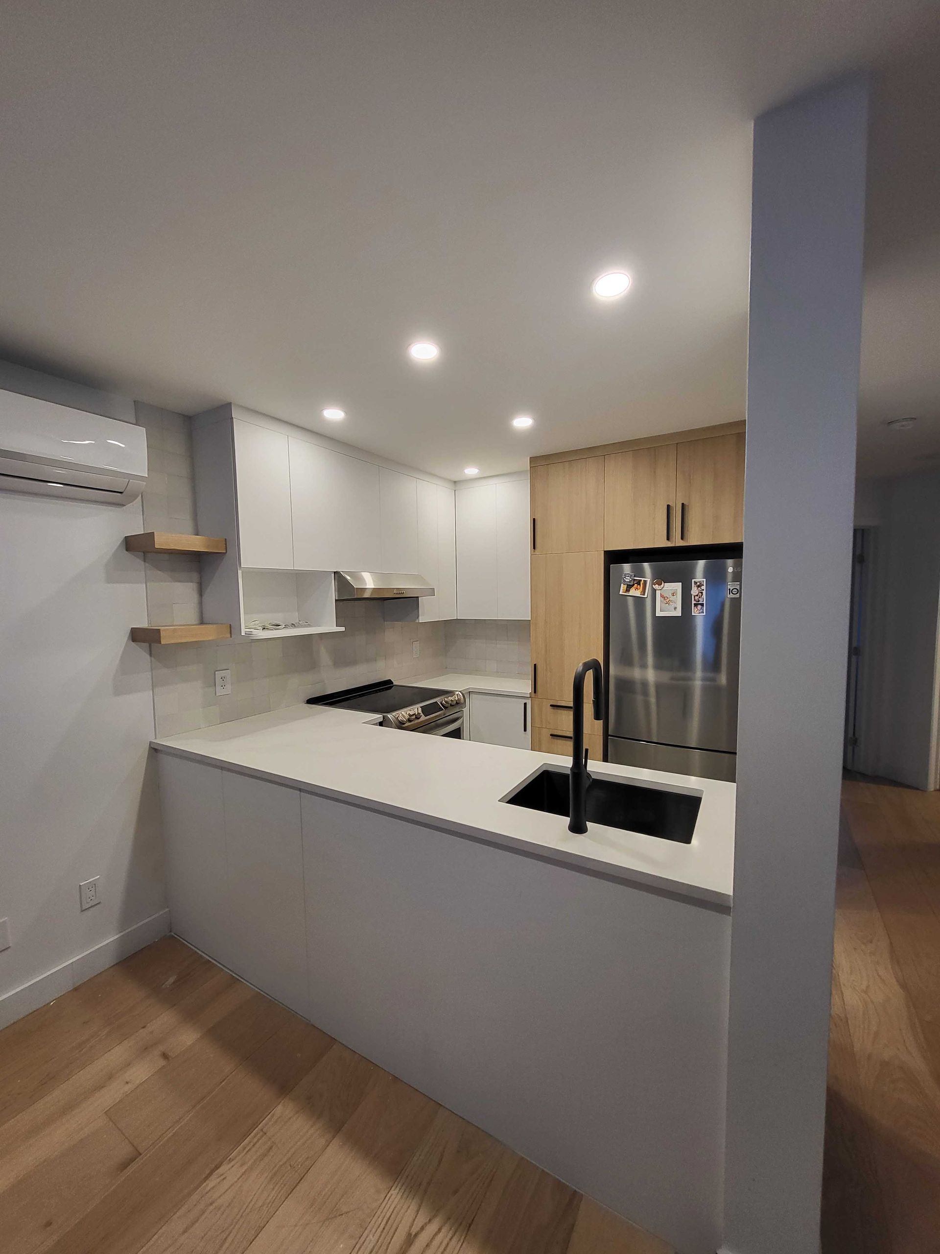 Cuisine moderne blanche avec îlot central, parquet, spots encastrés et réfrigérateur en acier inoxydable.
