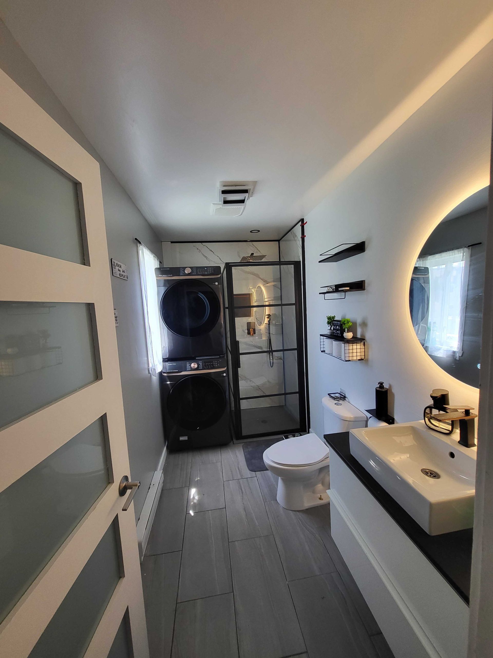 Salle de bains moderne et étroite avec meuble-lavabo blanc, miroir rond, porte de douche dépolie et laveuse-sécheuse superposées au fond.