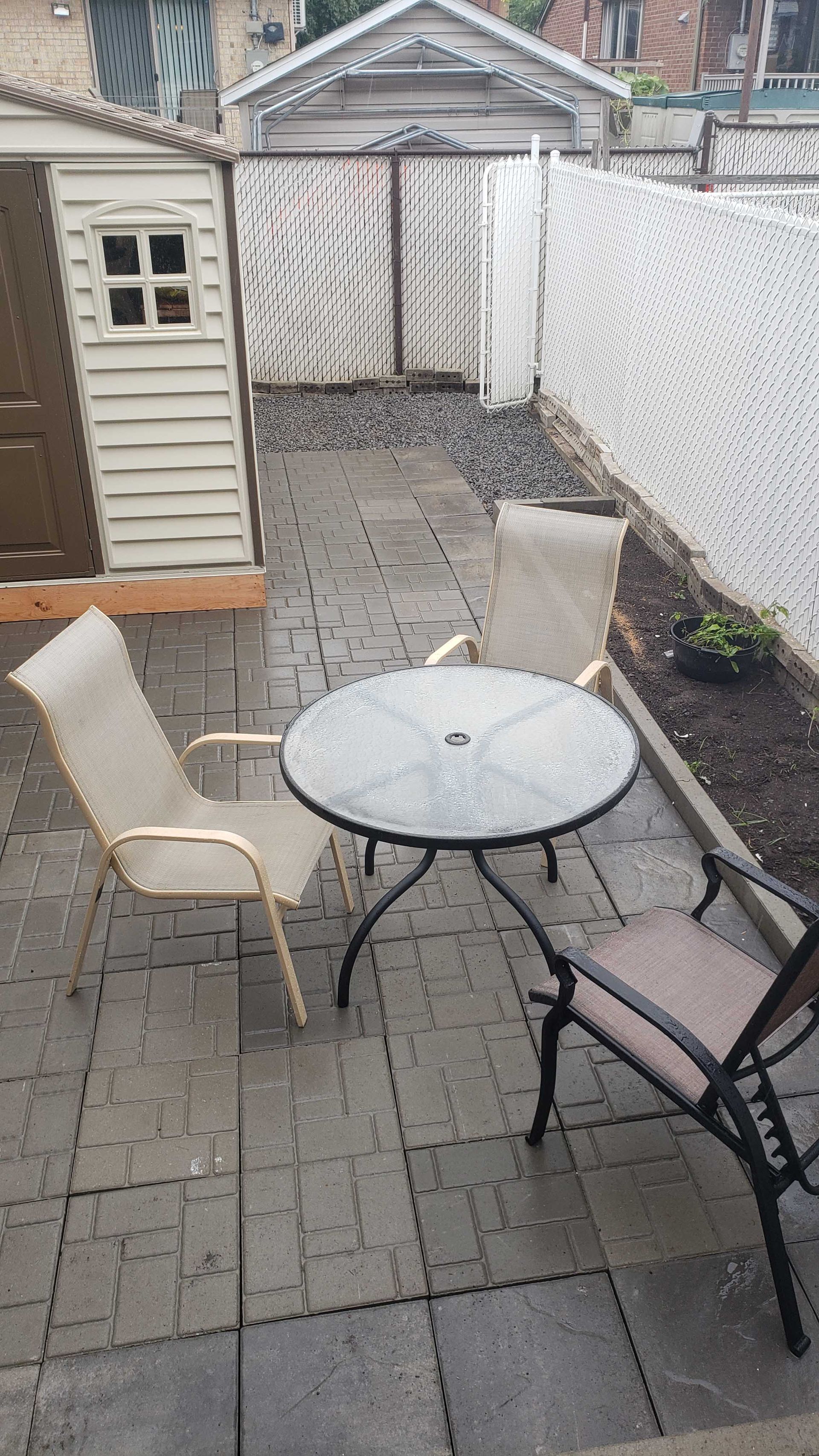 Petit patio avec table ronde, trois chaises, un abri de jardin et une clôture blanche, pavé de briques grises.