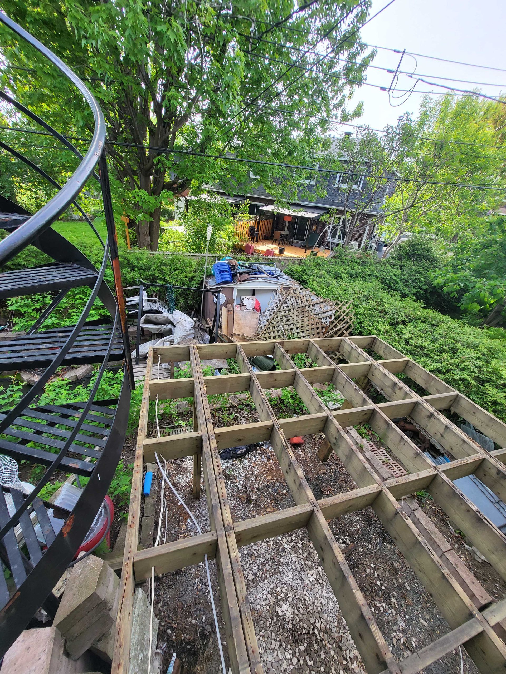 Cour arrière avec terrasse en bois, escalier en colimaçon et végétation luxuriante composée d'arbres et d'arbustes.