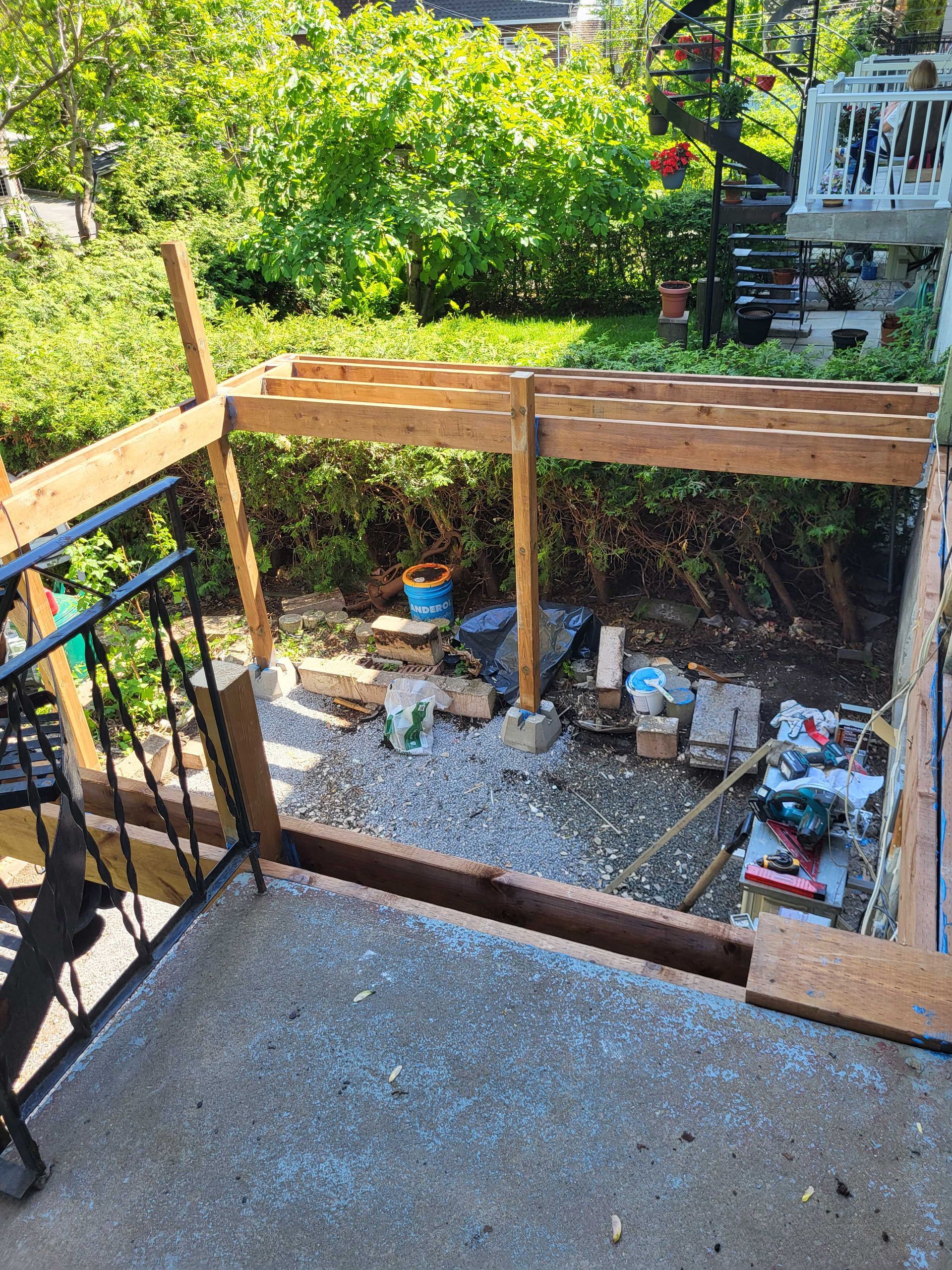 Terrasse en construction dans le jardin avec poteaux en bois, sol en gravier et arbres verts au-delà