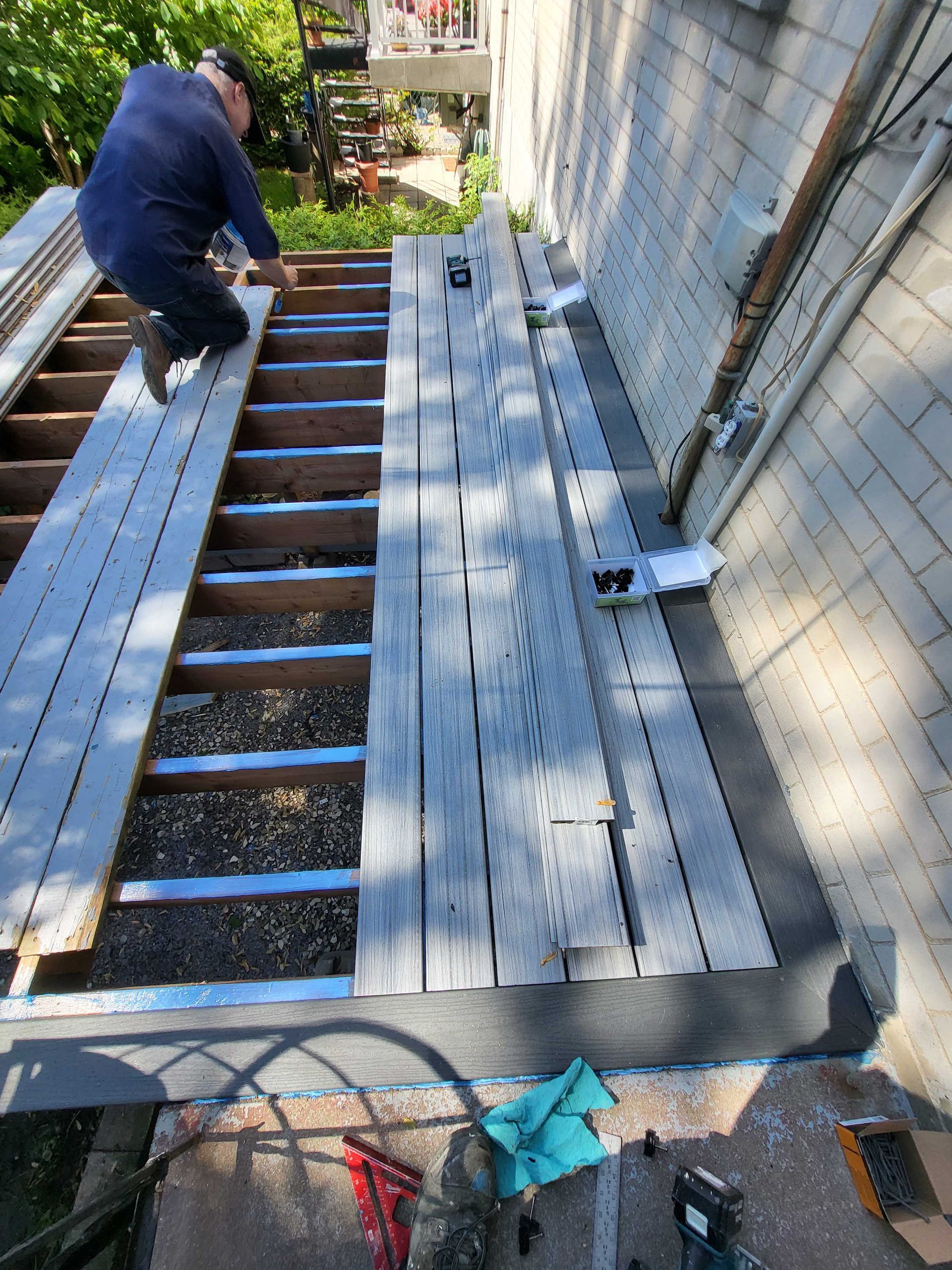 Un ouvrier installe des planches de terrasse grises le long d'un mur de briques, avec des solives apparentes et des outils à proximité.