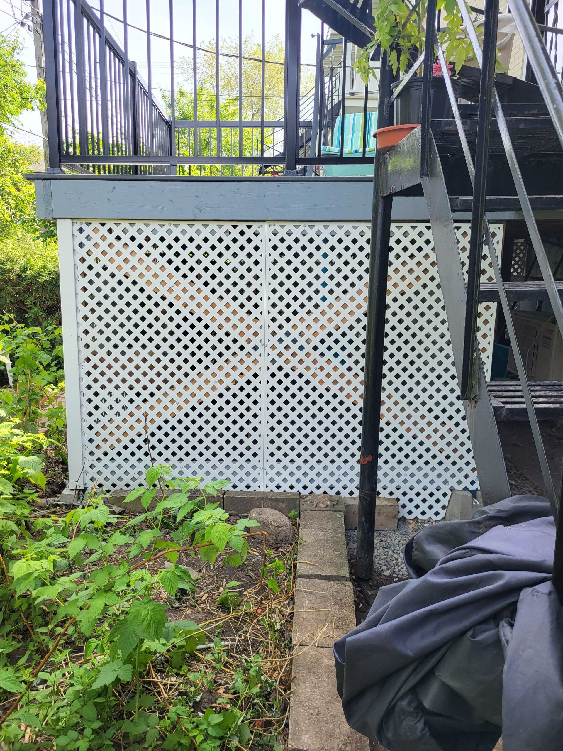 Un treillis blanc recouvre une terrasse avec des balustrades et des escaliers noirs, entourée de verdure et d'un sac noir.