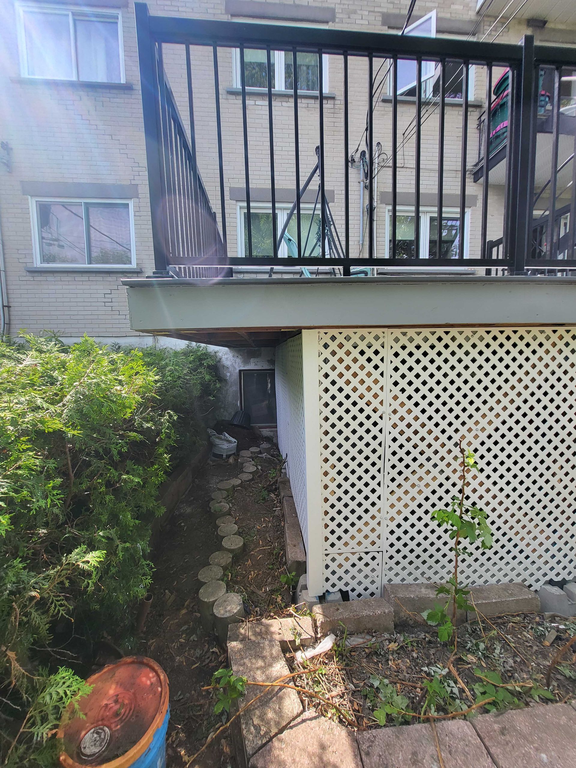Cour arrière sous une terrasse surélevée avec treillis blanc, chemin de pierre, arbustes et un pot bleu au premier plan