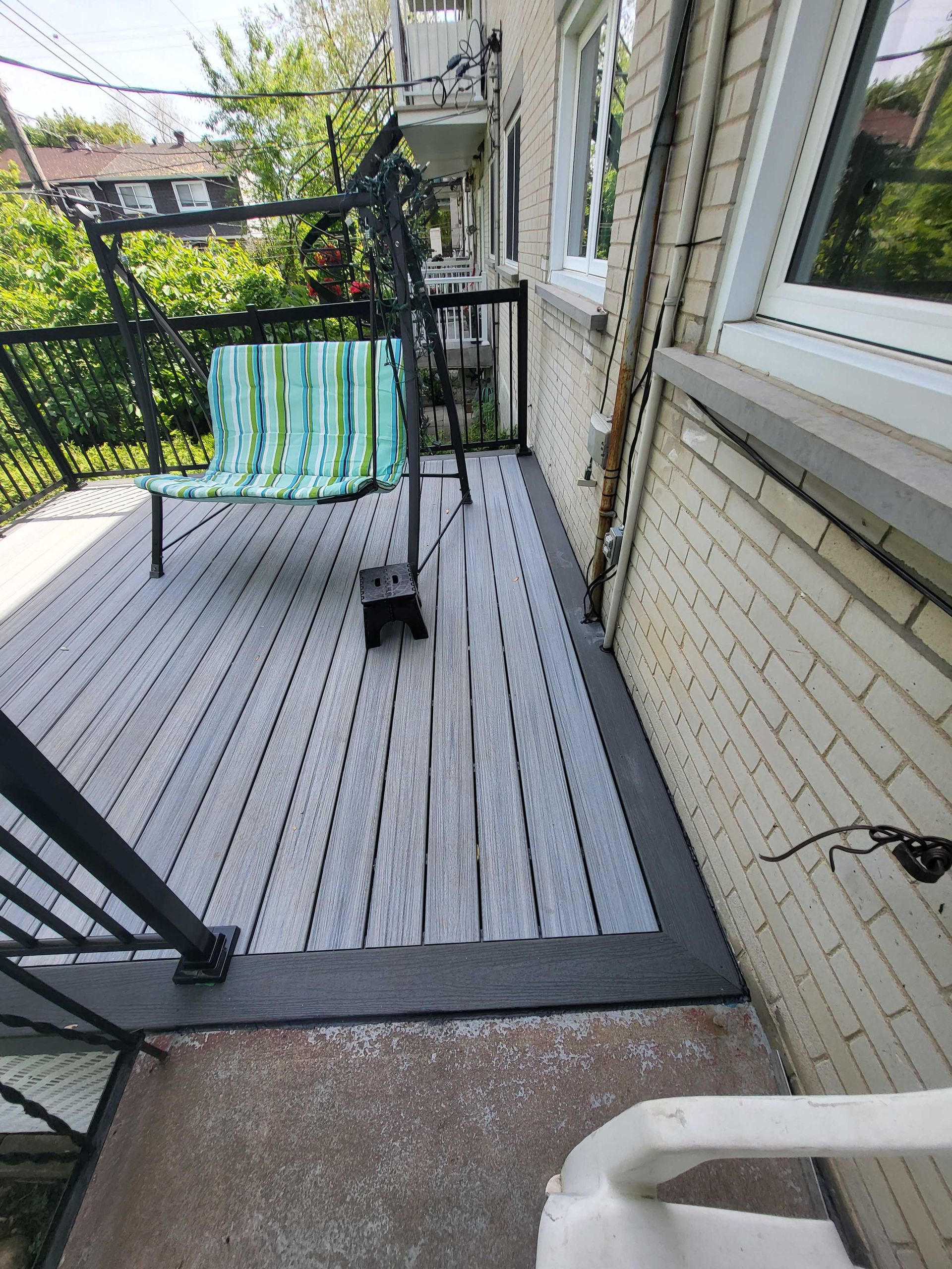 Petite terrasse dans le jardin avec chaise rayée, balustrade noire et mur de maison en briques blanches.