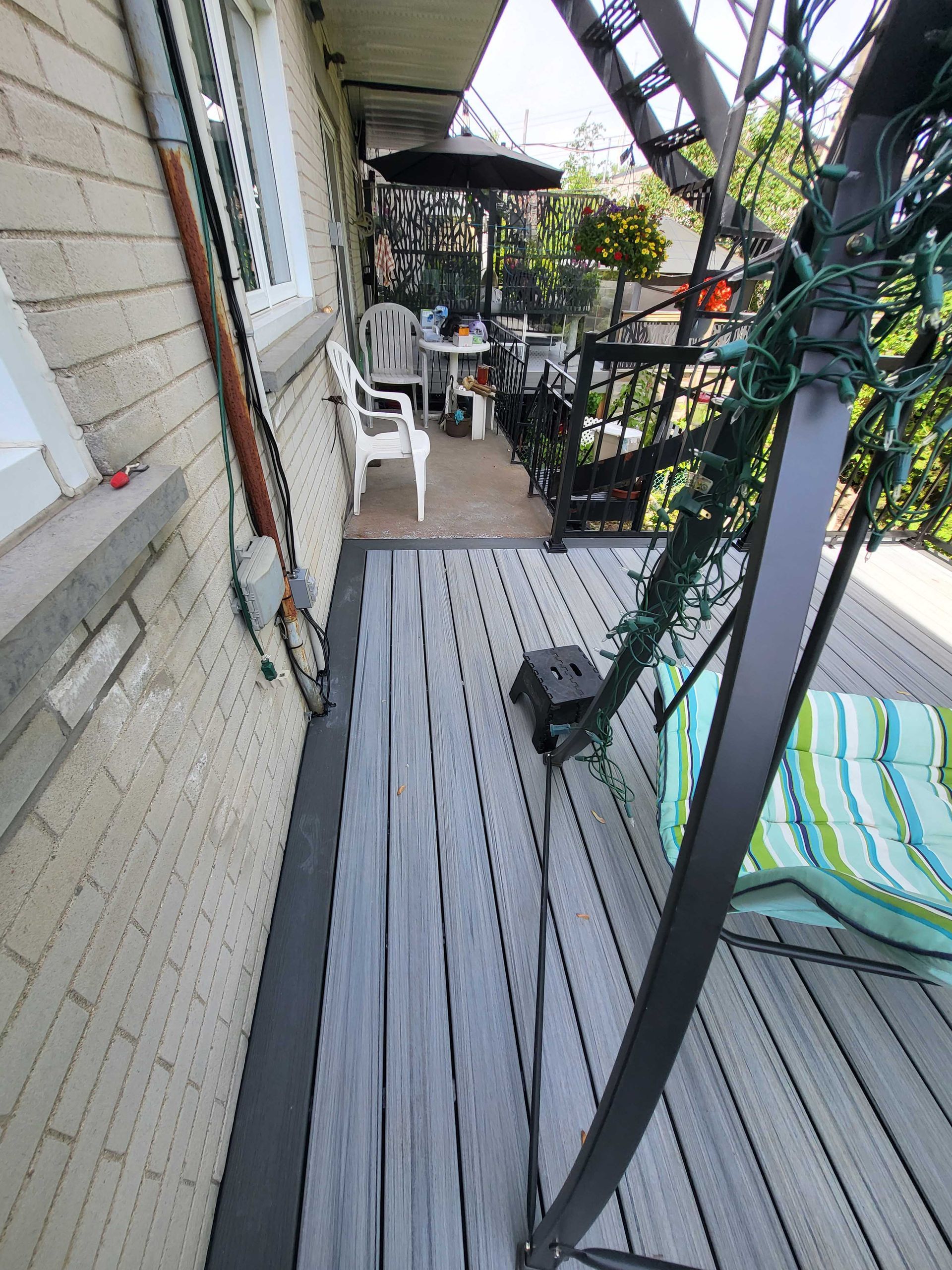 Étroite véranda en bois avec rambarde, plantes en pot et mobilier de jardin jouxtant la façade d'une maison.