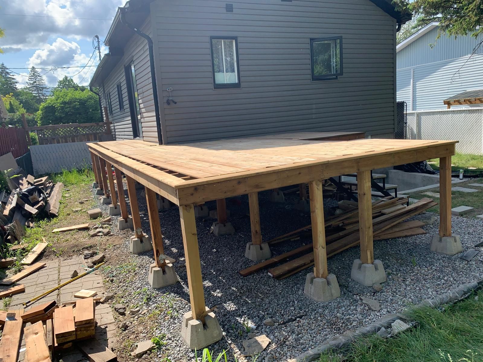 Terrasse en bois surélevée en construction à côté d'une maison grise, soutenue par des poteaux sur des fondations en béton.
