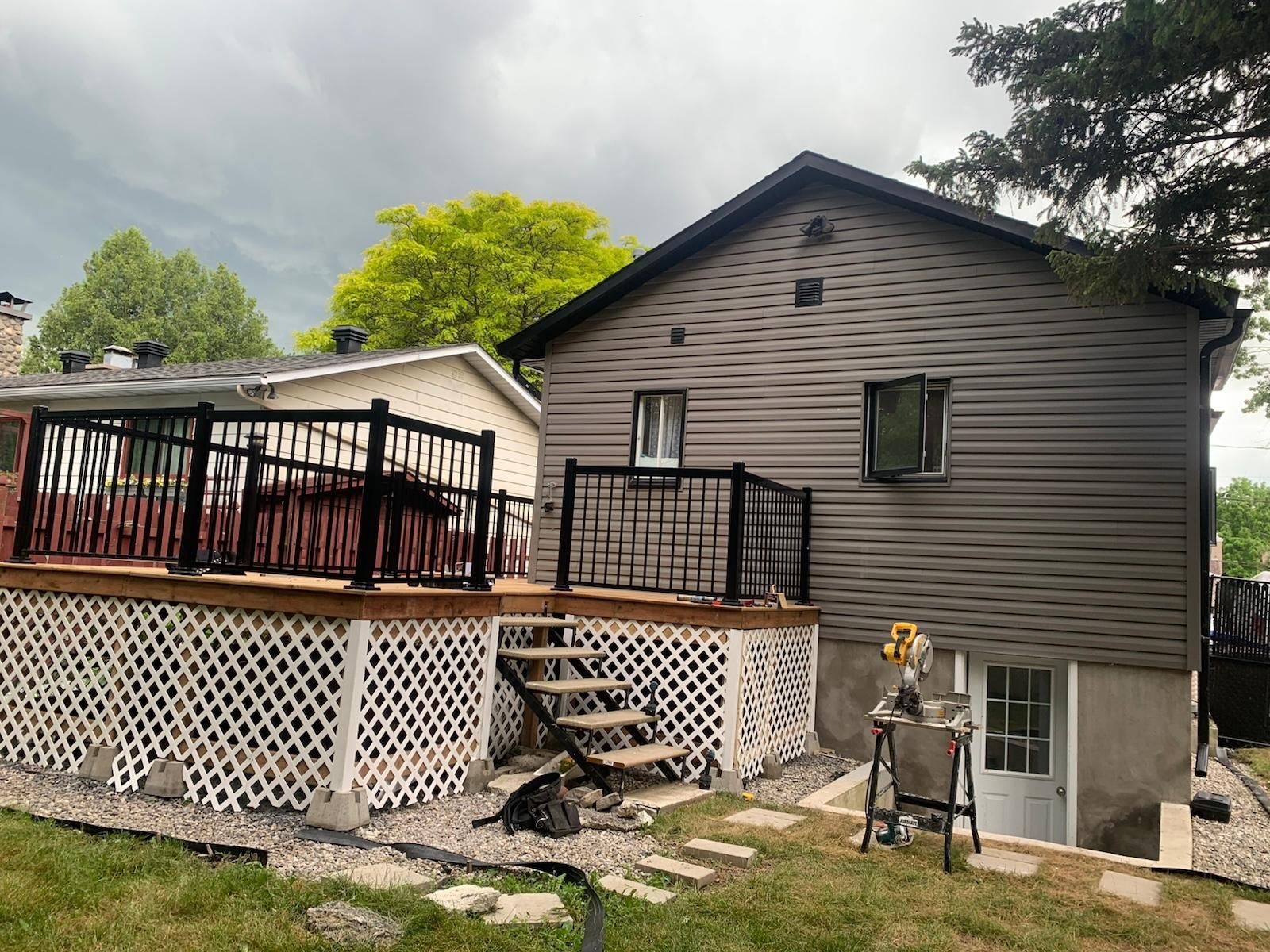Terrasse arrière et maison aux murs gris avec escalier, plinthe en treillis et mobilier de patio sous un ciel nuageux