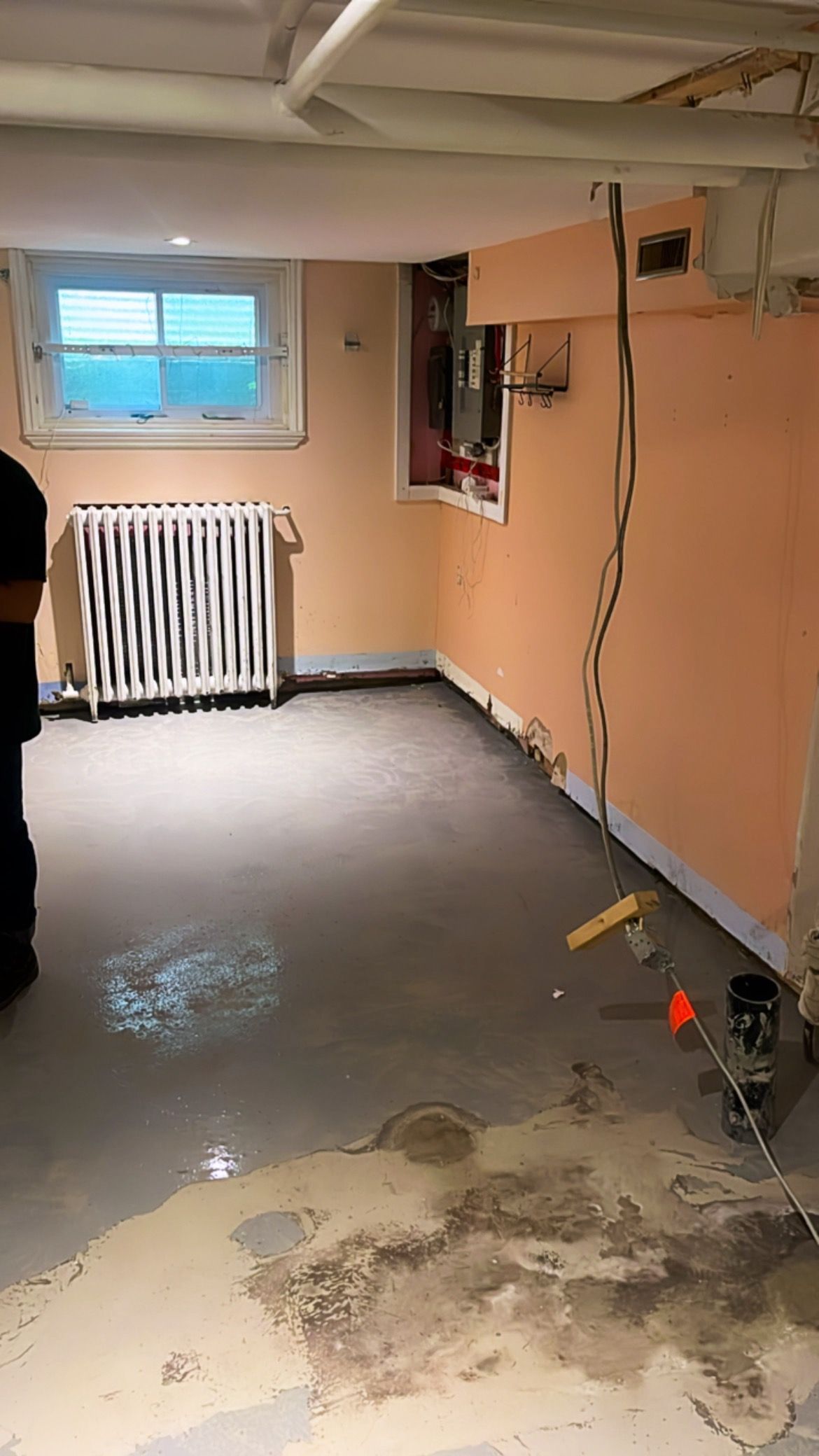 Pièce au sous-sol avec des murs couleur pêche non finis, une petite fenêtre, des fils électriques apparents et des taches d'humidité sur le sol.