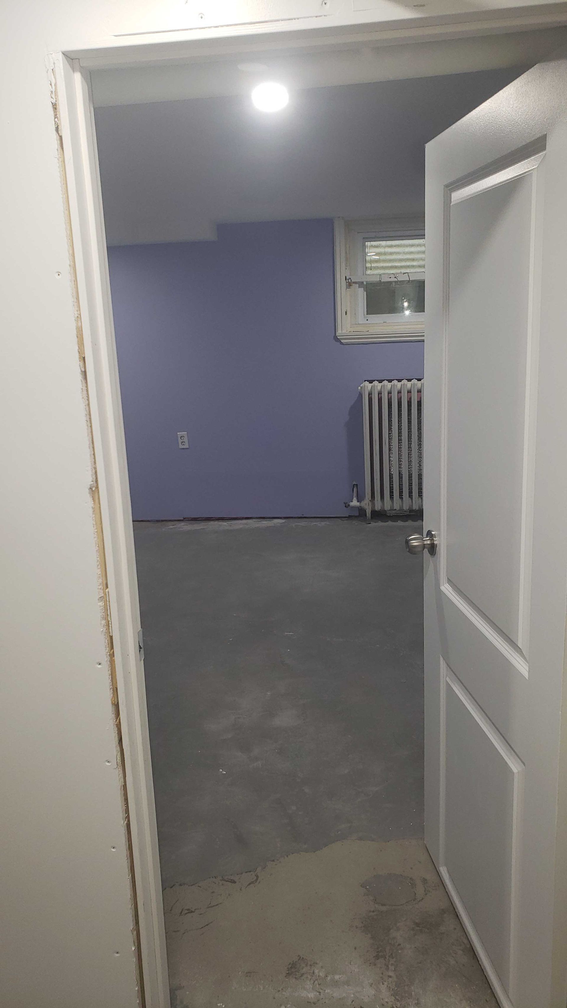 Porte ouverte donnant sur une pièce vide aux murs violets, à la moquette grise et avec un radiateur sous une fenêtre.