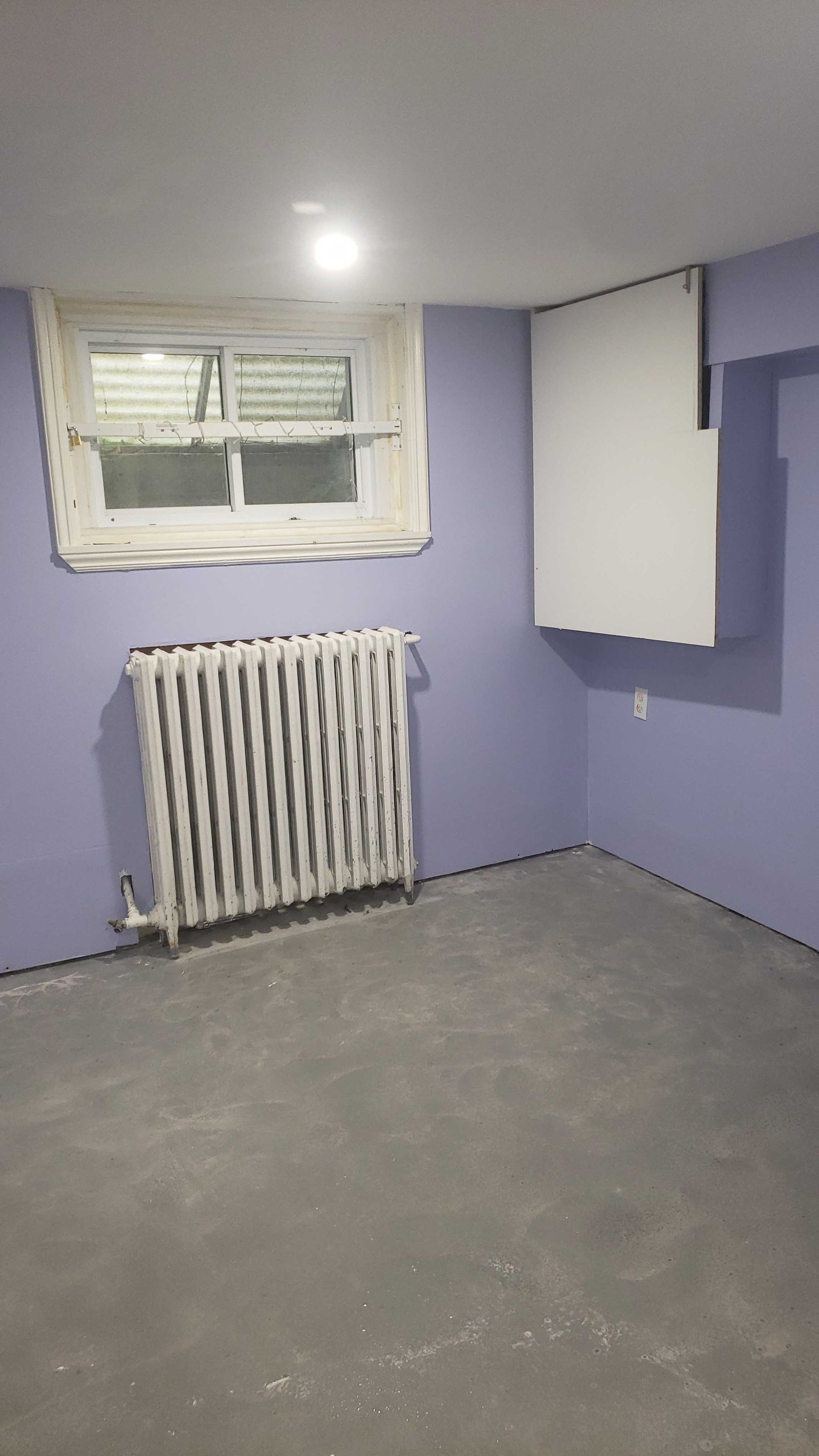 Pièce violette vide avec un radiateur, une petite fenêtre et une armoire blanche au-dessus.