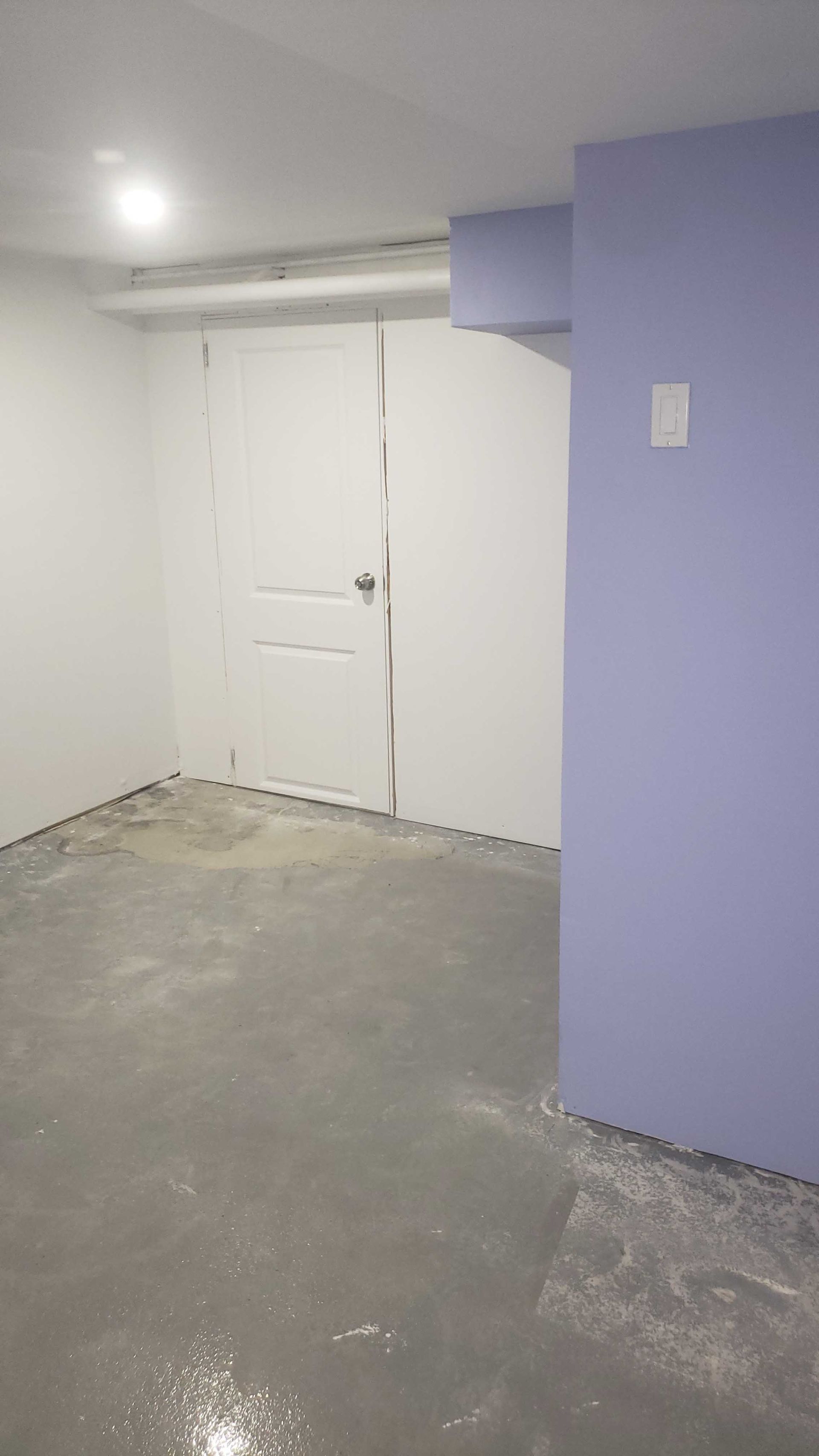 Pièce vide et inachevée avec des portes doubles blanches, un sol en béton et un mur lavande.