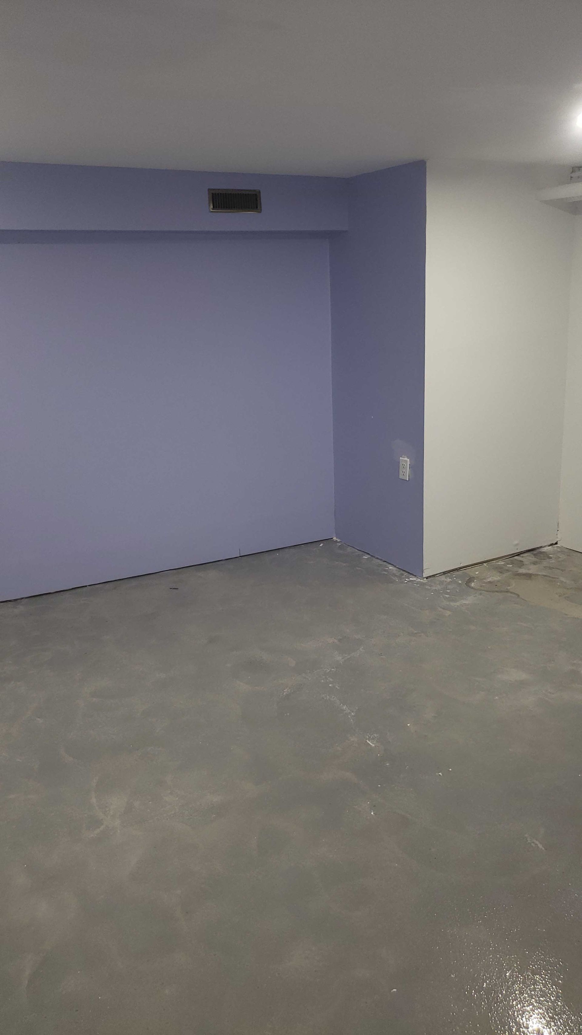 Pièce vide et inachevée avec des murs violets, un sol en béton gris et un mur d'angle blanc.