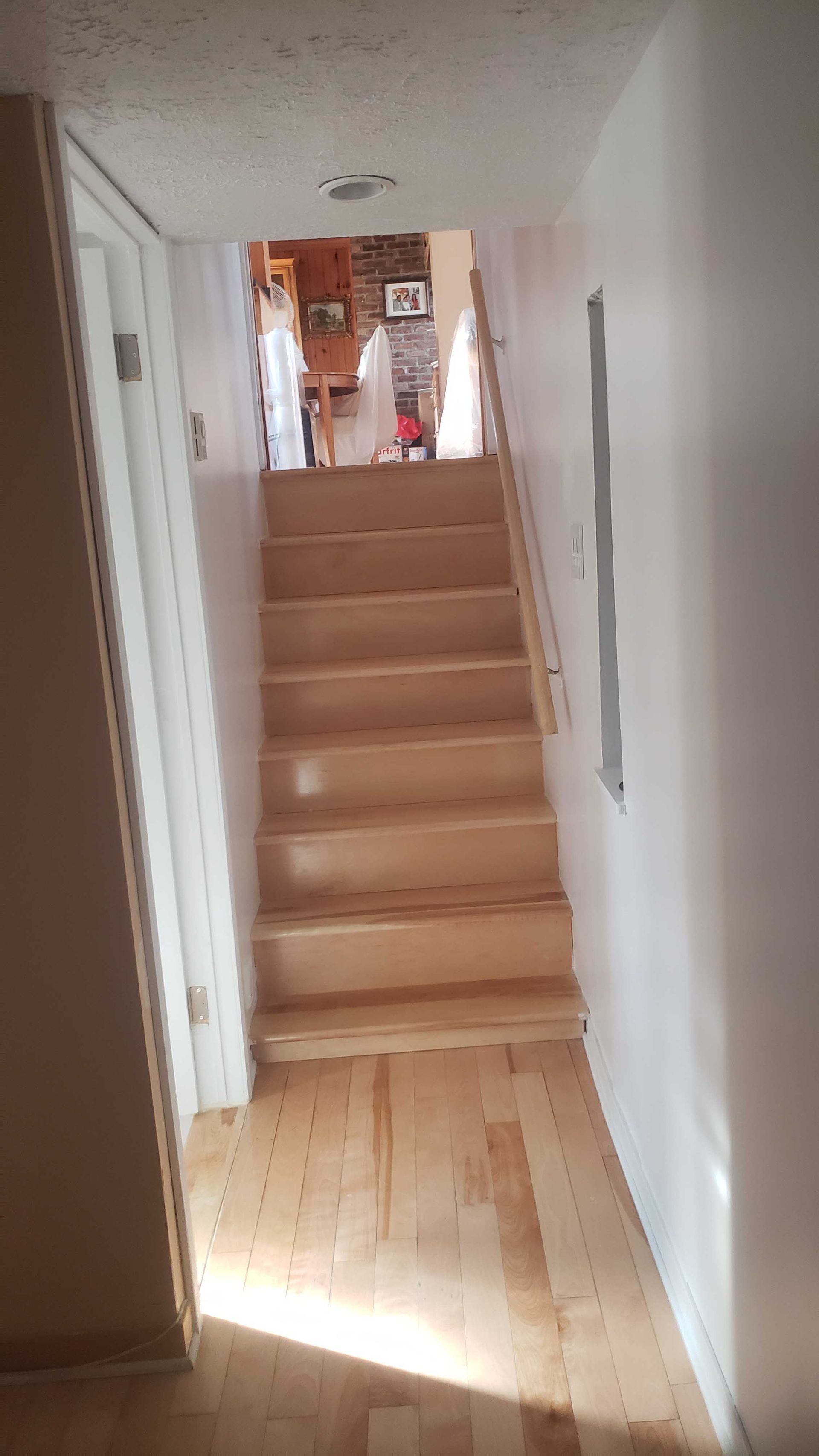 Couloir étroit avec un escalier en bois clair menant à l'étage, murs blancs et lumière du soleil sur le sol.