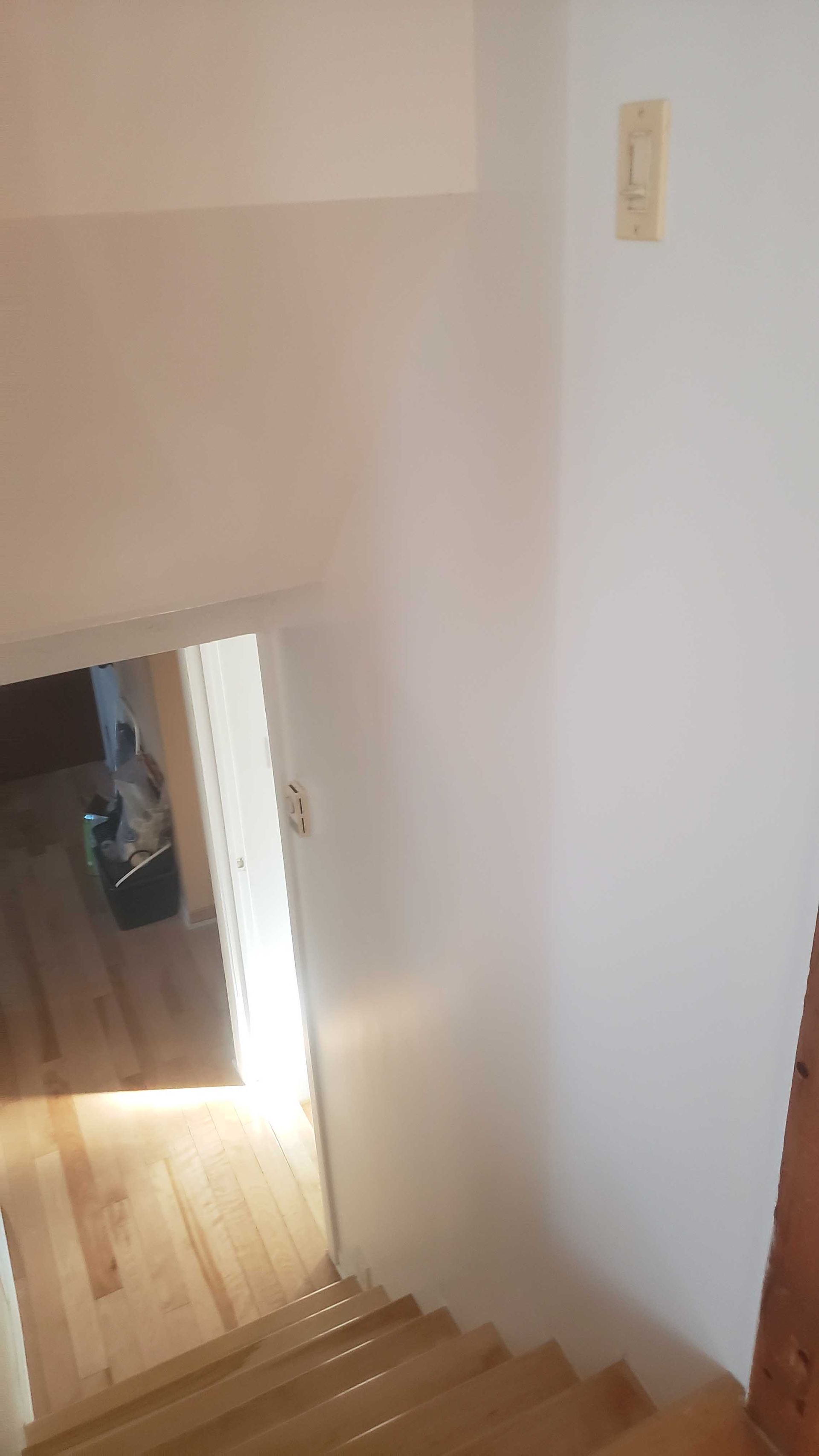 Cage d'escalier aux murs blancs, marches en bois et une porte en contrebas laissant entrer la lumière du soleil