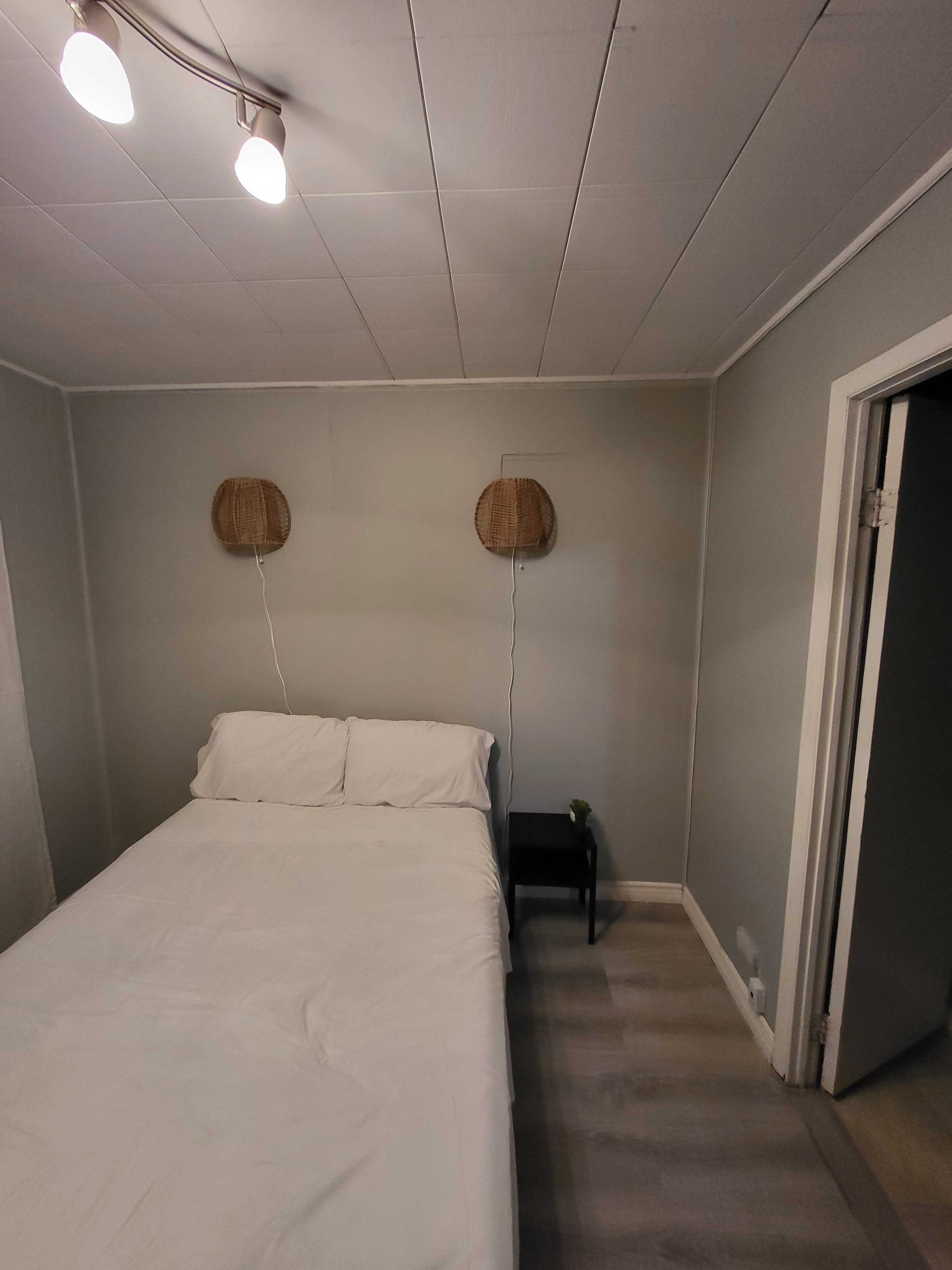 Petite chambre grise avec un lit blanc, deux paniers muraux et une porte ouverte.