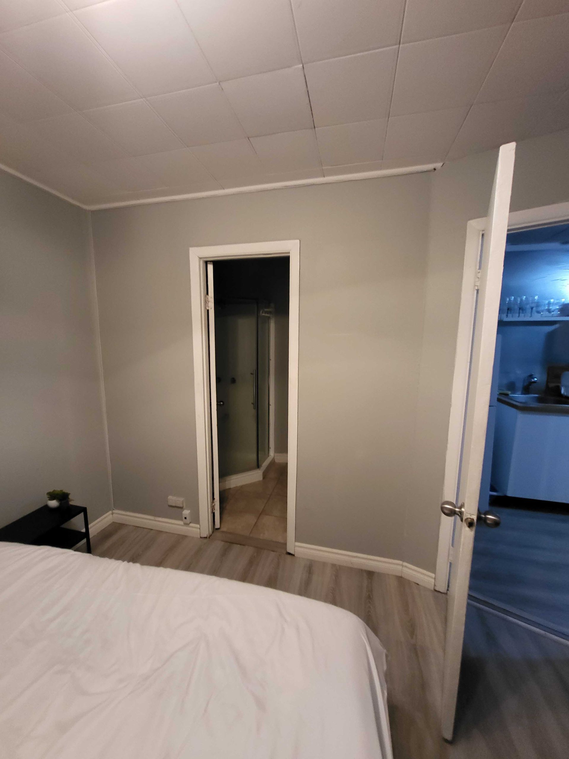 Chambre aux murs gris, lit blanc, porte ouverte sur un couloir et porte bleue entrouverte.