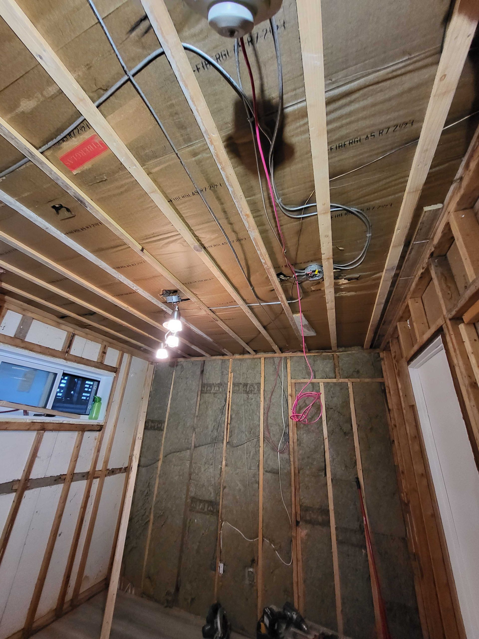 Intérieur d'une pièce partiellement aménagée avec poutres apparentes au plafond, isolation, suspension lumineuse et murs non finis.
