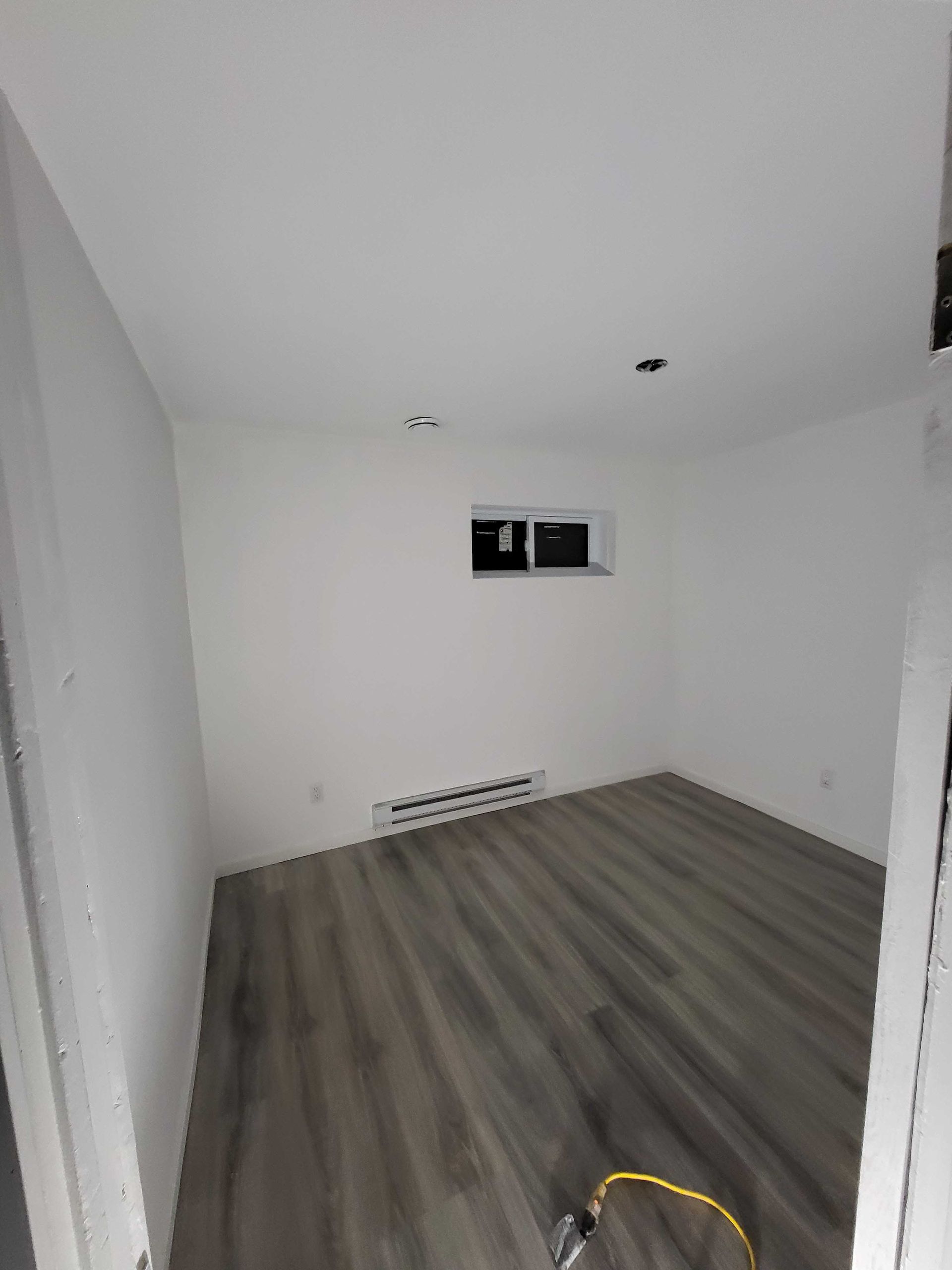 Pièce blanche vide avec un parquet gris et une petite fenêtre sur le mur du fond.