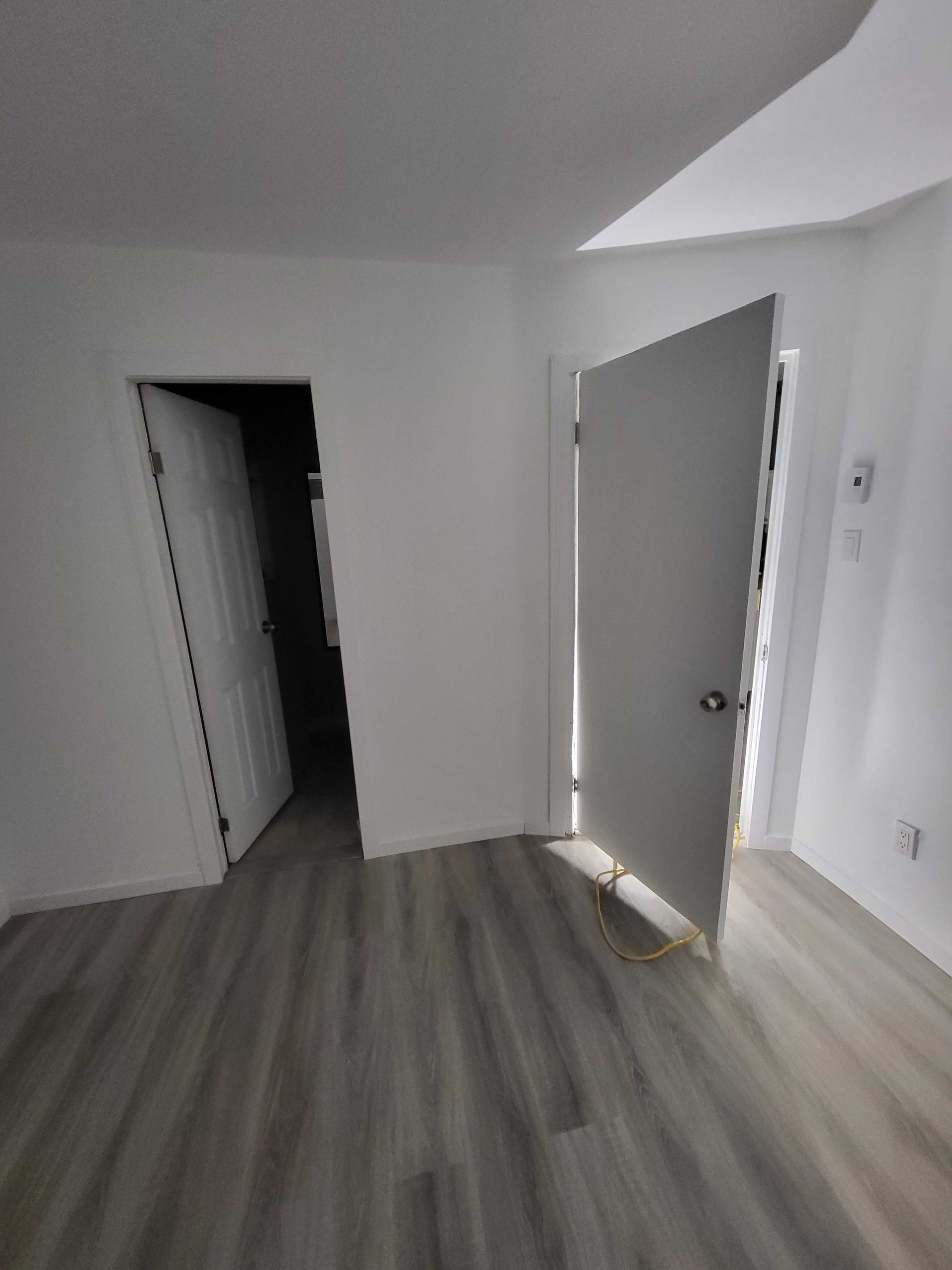 Pièce blanche vide avec parquet gris, porte ouverte et grande fenêtre laissant entrer la lumière du soleil.